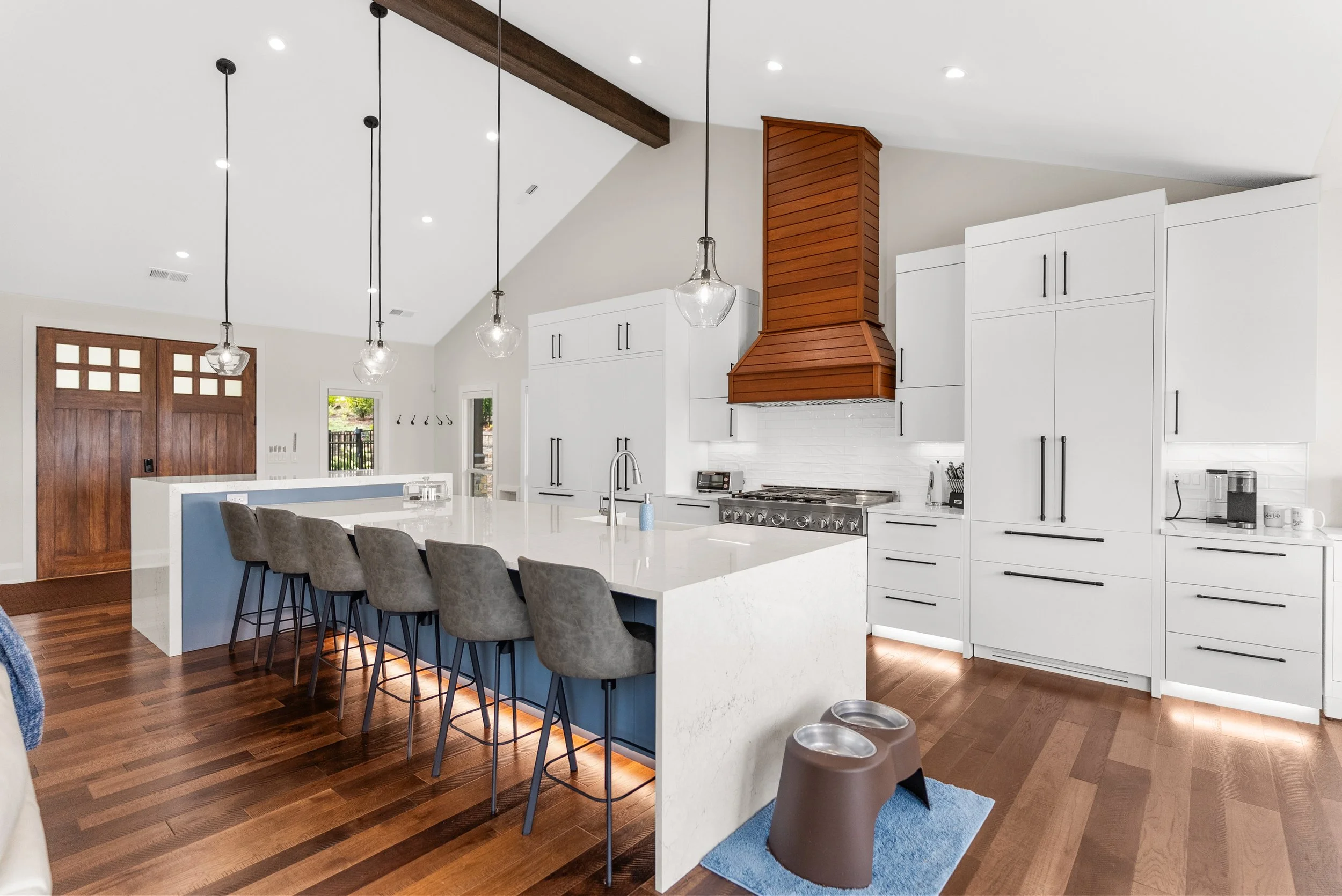 Modern kitchen featuring white cabinets, a large island with a white countertop, and a wooden range hood over a stainless steel stove. Pendant lights hang above the island. There's a front door with wooden panels and glass, and a small window near th