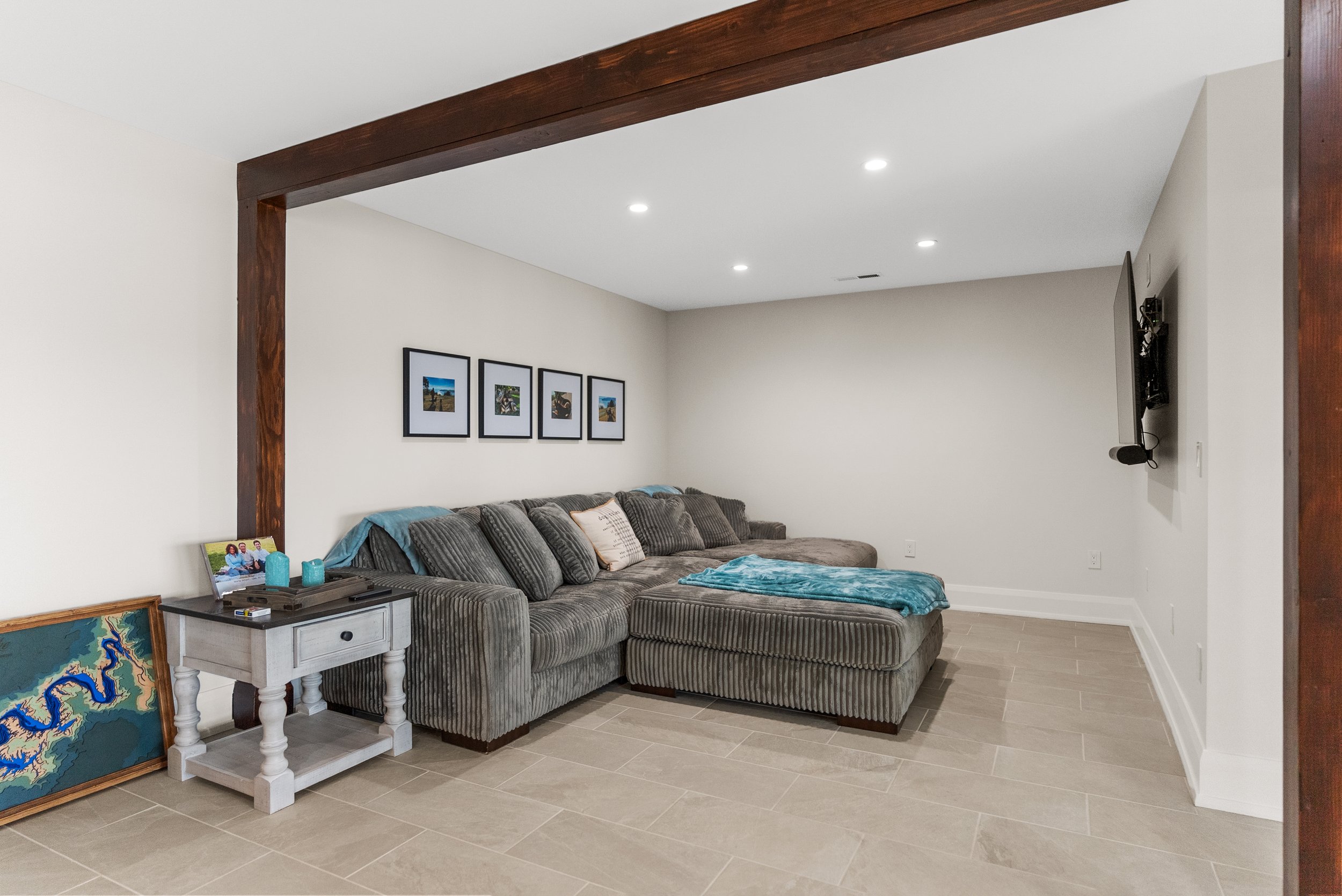 Living room with gray sectional sofa, blue blanket, framed pictures on wall, white side table with family photo and candles, wall-mounted TV with microphone, light gray tiled floor, white walls, and wooden beam in ceiling.