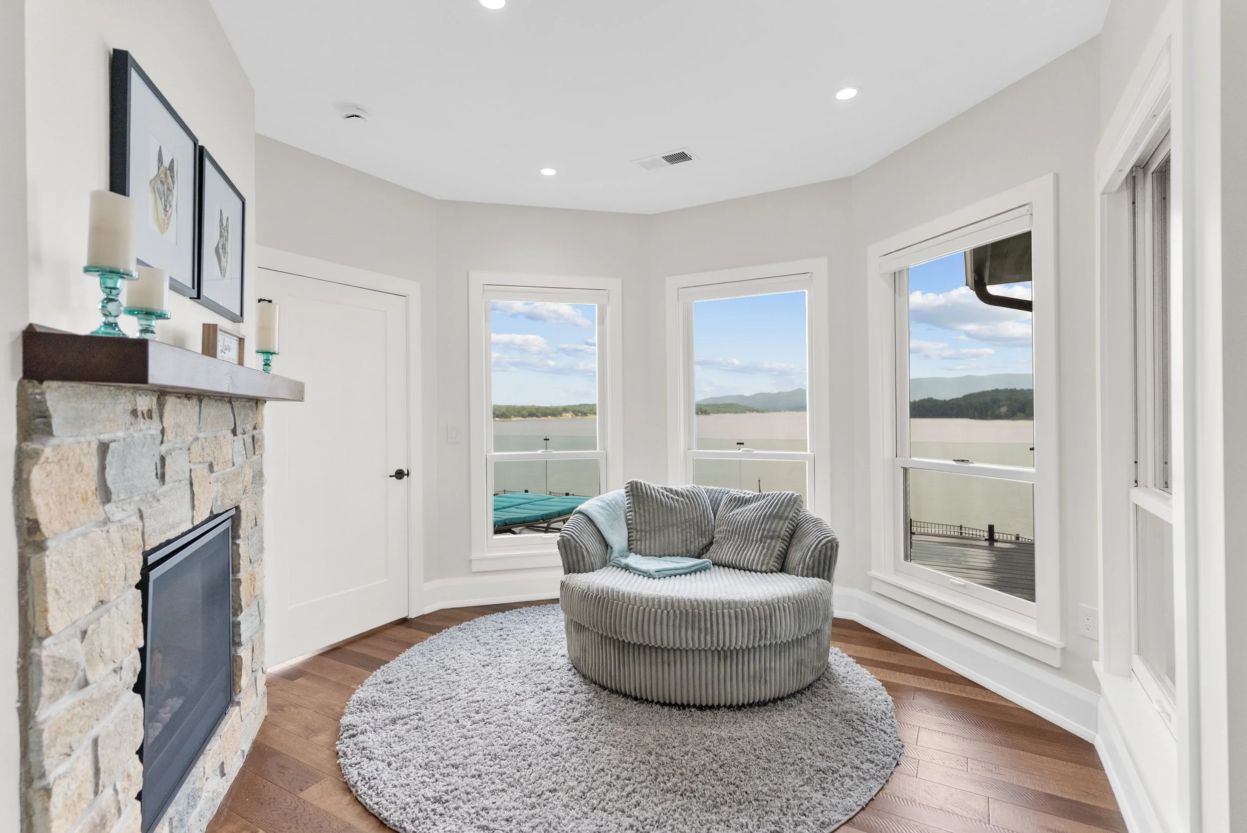 A cozy living room with large windows overlooking a lake and mountains, featuring a round striped armchair, a stone fireplace with a wooden mantel decorated with candles, and a light-colored round rug.