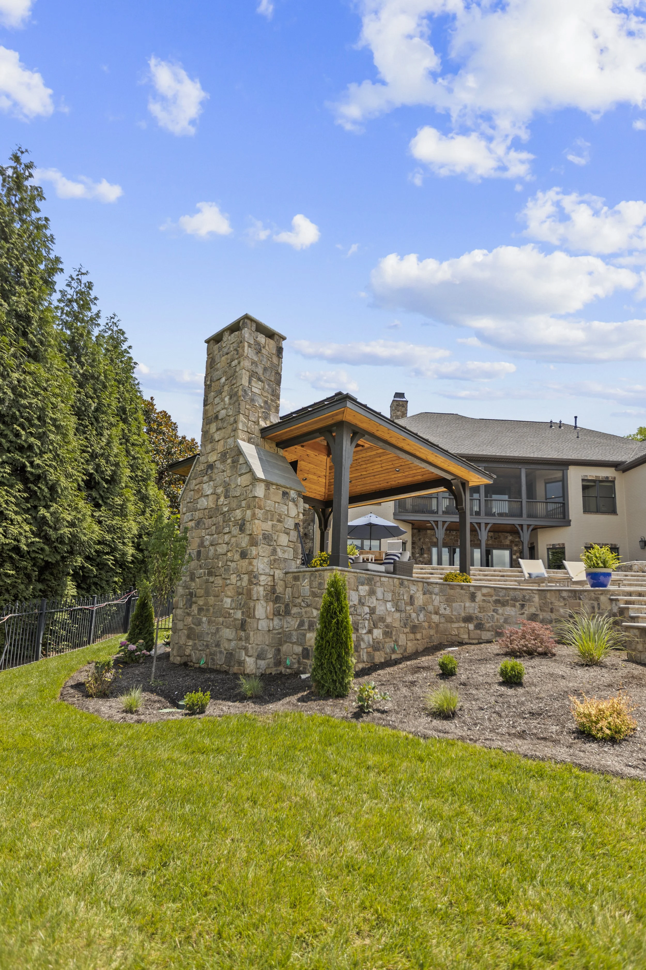A backyard with a stone chimney, covered patio area with furniture, and a green lawn with landscaped plants, neighboring house is visible in the background under a partly cloudy sky.