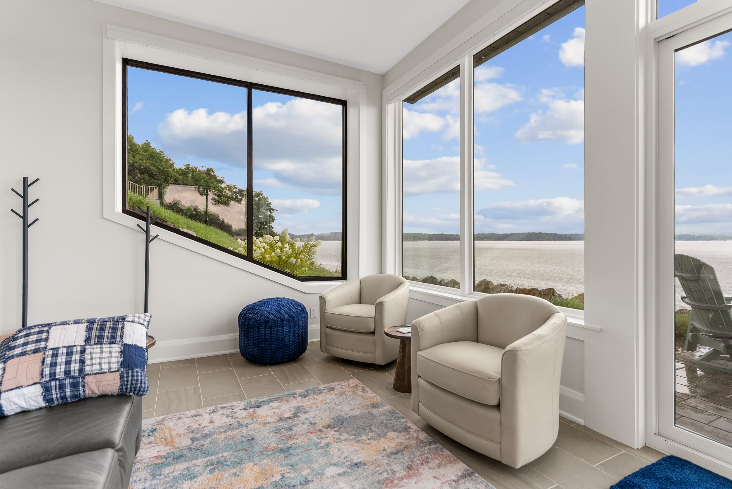 Living room with large windows overlooking a lake, featuring white chairs, a colorful rug, and a patchwork quilt on a sofa.