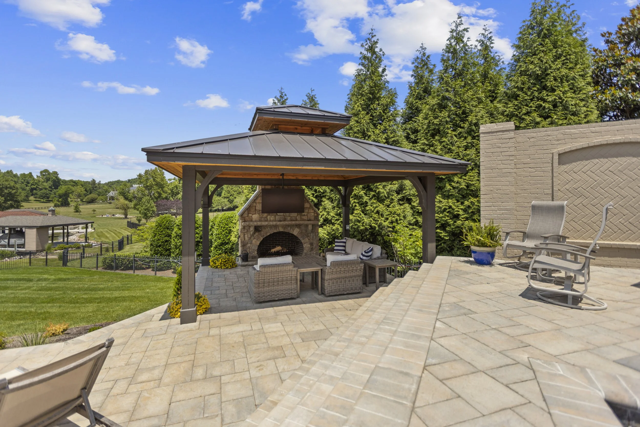 Patio with outdoor fireplace, seating area, and a grill, overlooking a green yard with trees and a fence under a blue sky with clouds.