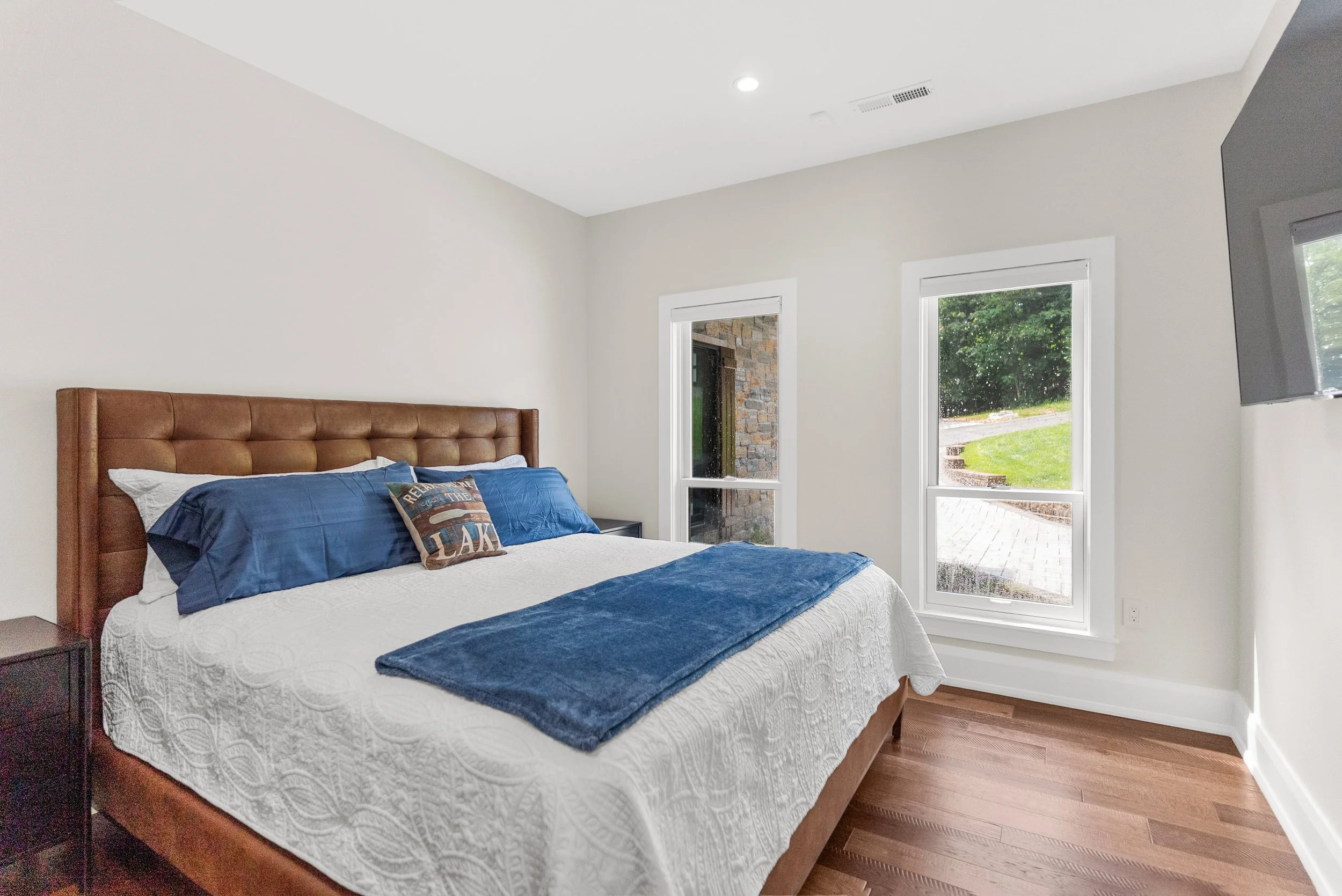 Bedroom with a tall wooden bed frame, white quilted bedspread, blue bed runner, matching blue pillows, window with view of greenery and a brick patio, and a wall-mounted TV.