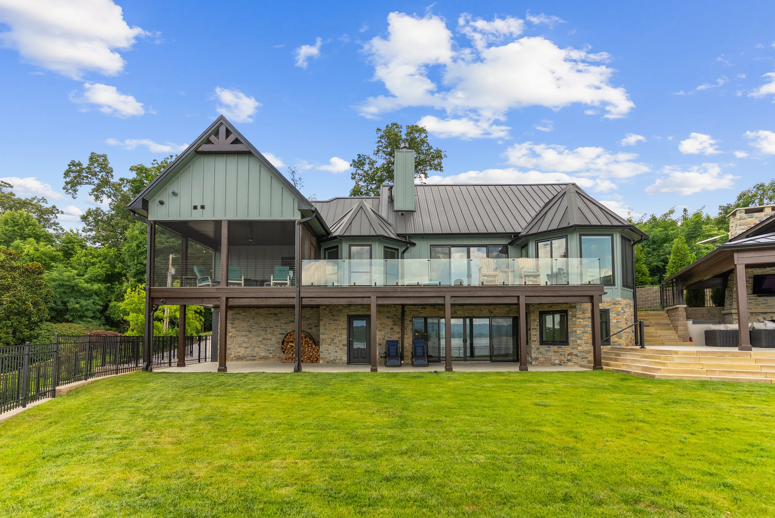 Backyard view of a modern house with a stone foundation, green siding, and large glass windows, featuring a wooden deck with outdoor furniture, stairs, and a lawn, surrounded by trees under a partly cloudy sky.