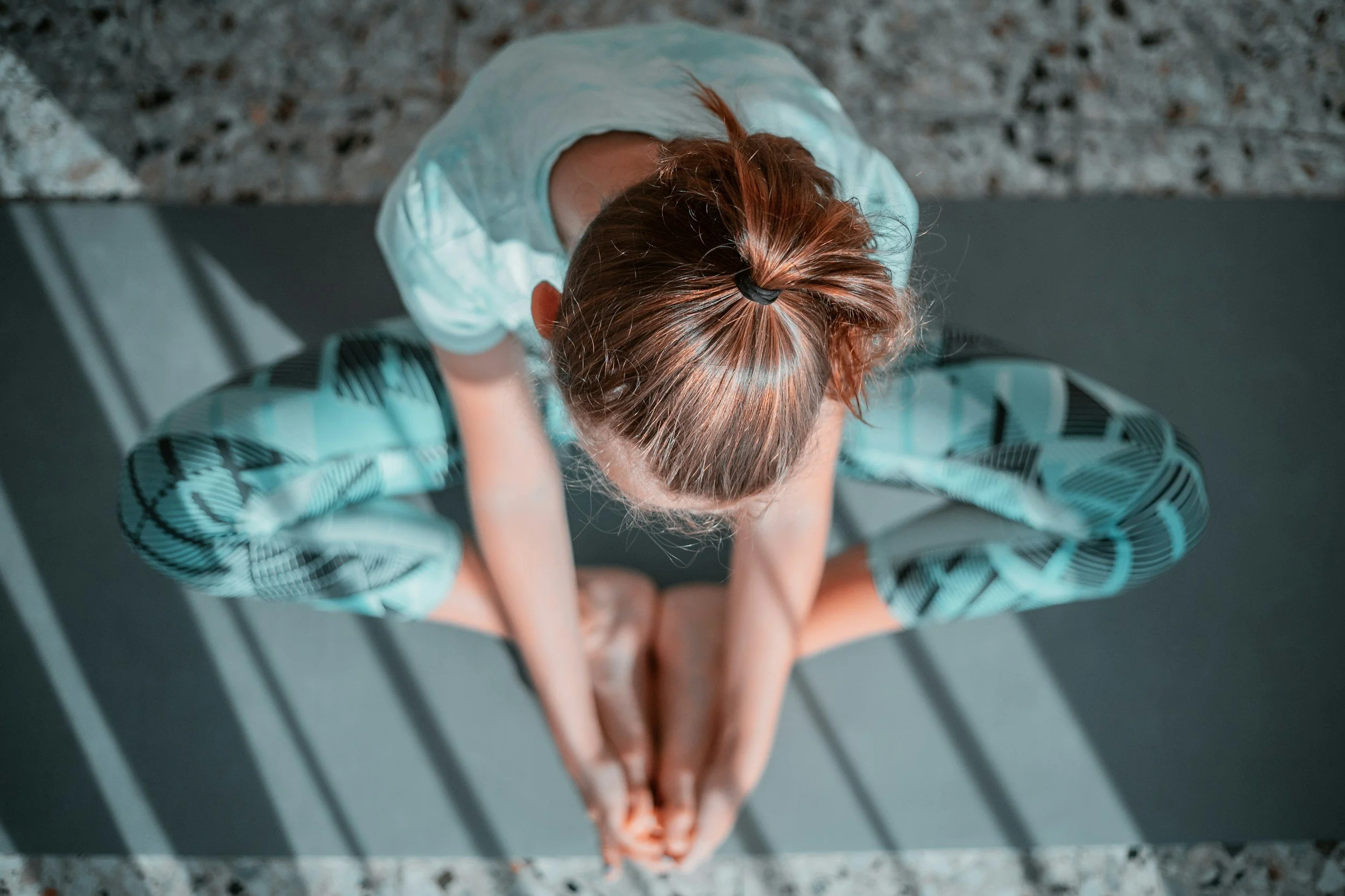 Overhead view of a child sitting on a bench with her head down, wearing a light blue jacket and patterned pants, on a terrazzo floor.