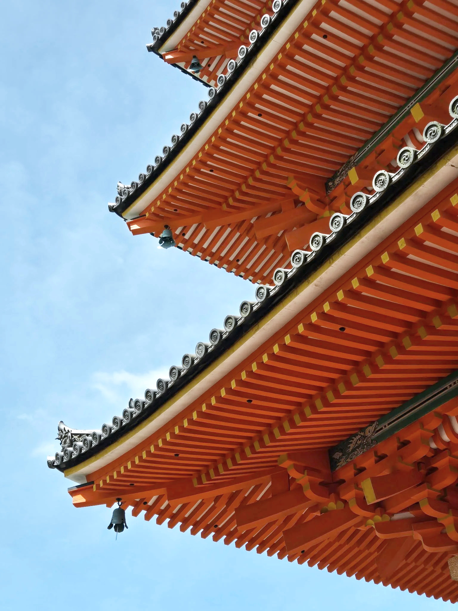Close-up of the corner of a traditional Asian temple roof with ornate decorative elements, including a small bell hanging from the corner, against a clear blue sky.