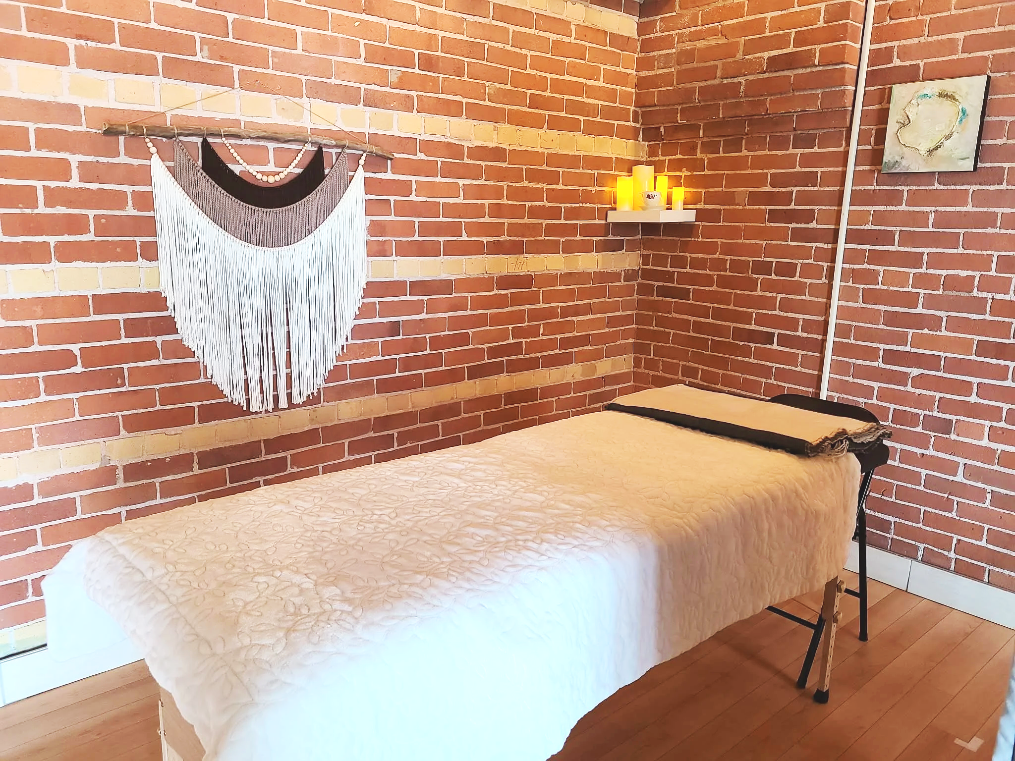 A cozy room with exposed brick walls, a massage table with a quilted beige bedspread, a small shelf holding candles, a piece of wall art, and a macramé wall hanging.
