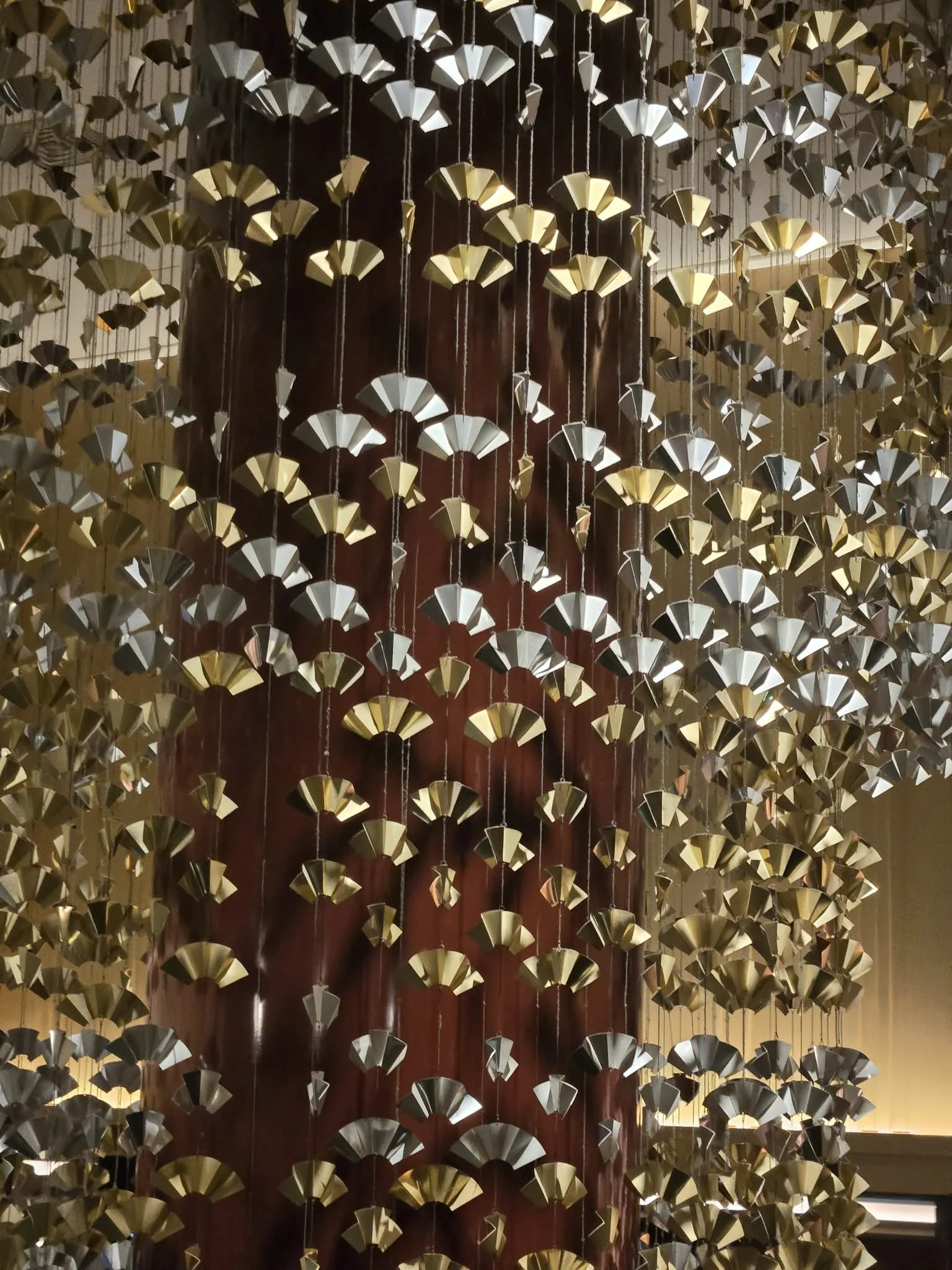 A decorative hanging installation made of numerous small paper fan-shaped elements, suspended by strings, creating a layered, cascading effect against a dark background.