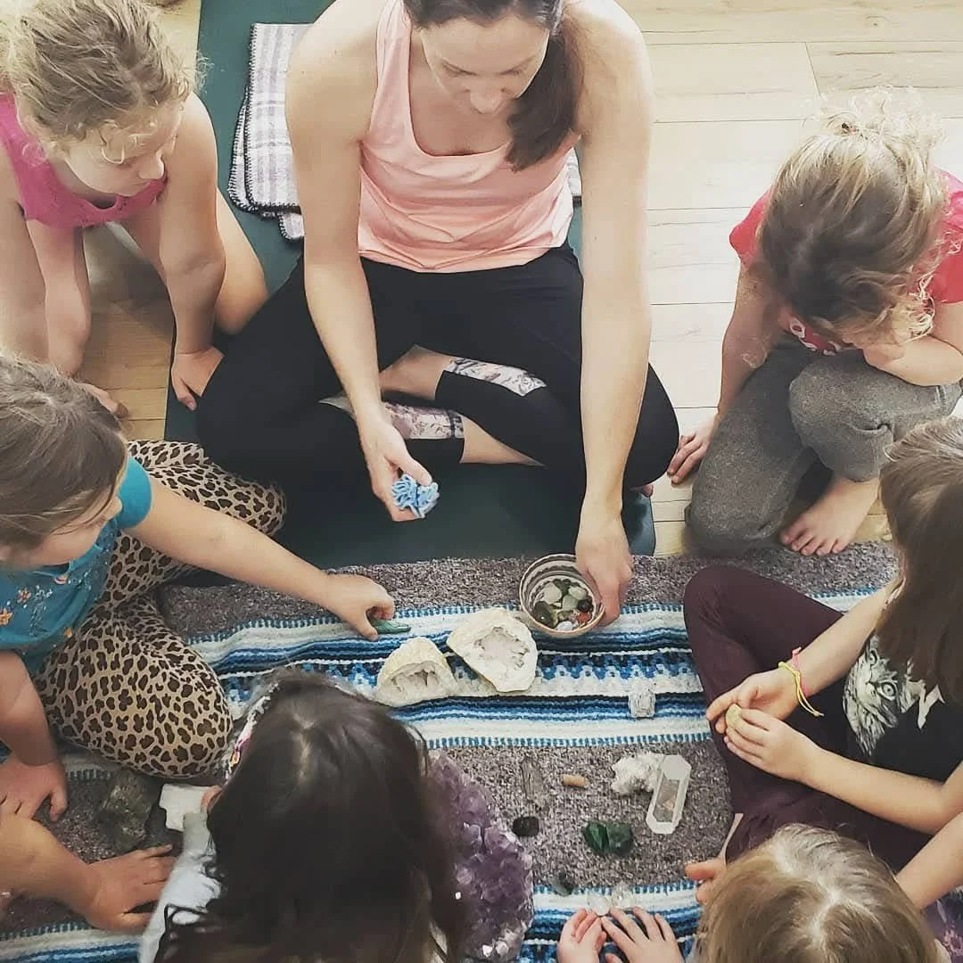 A woman leading a group of children in a meditation or mindfulness activity, looking and handling crystals, sitting cross-legged on a colourful rug in a room with wooden floors.