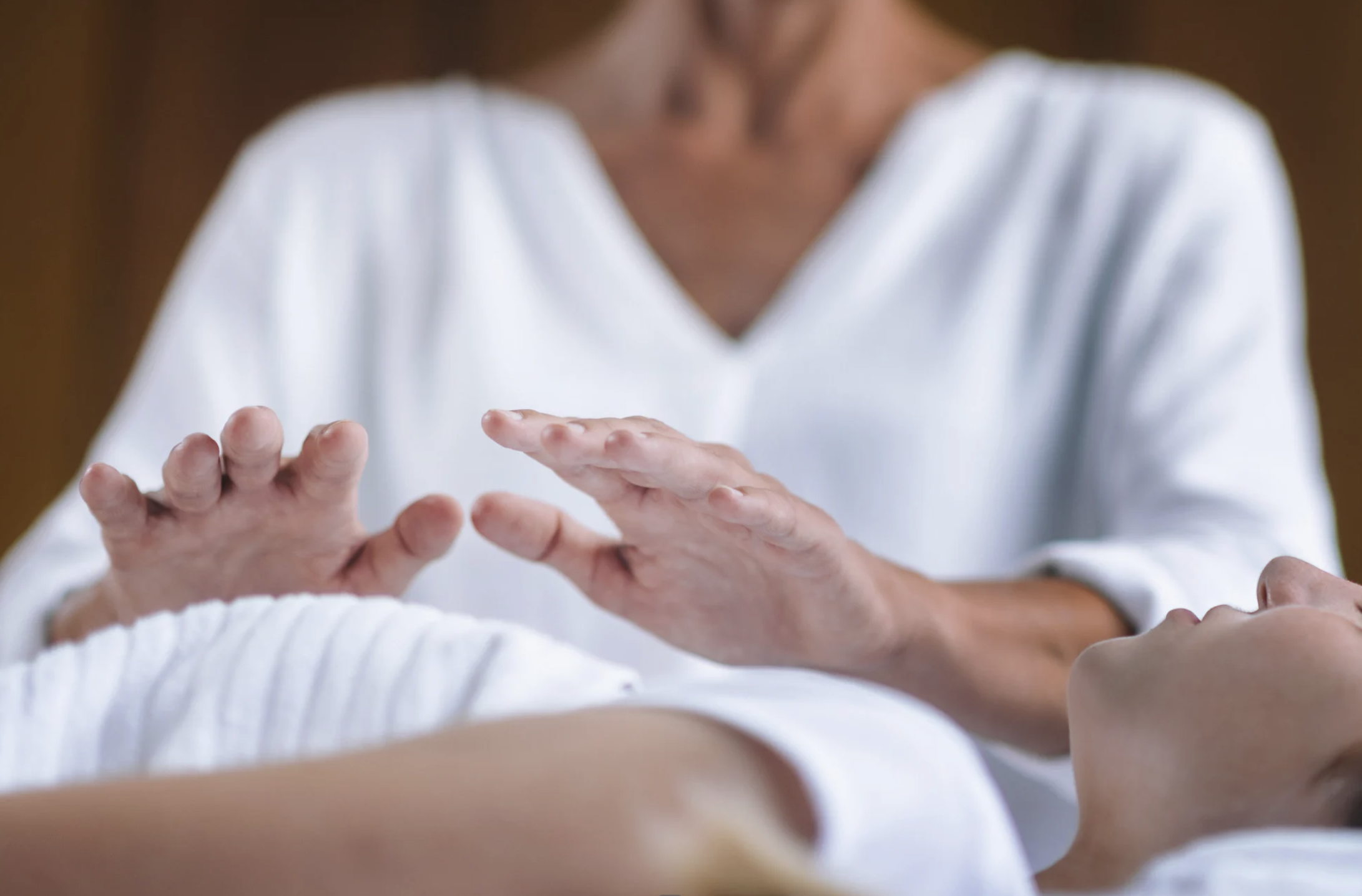 A Reiki practitioner in a white shirt gently holds their hands above a client lying on a treatment table in a calm indoor setting.
