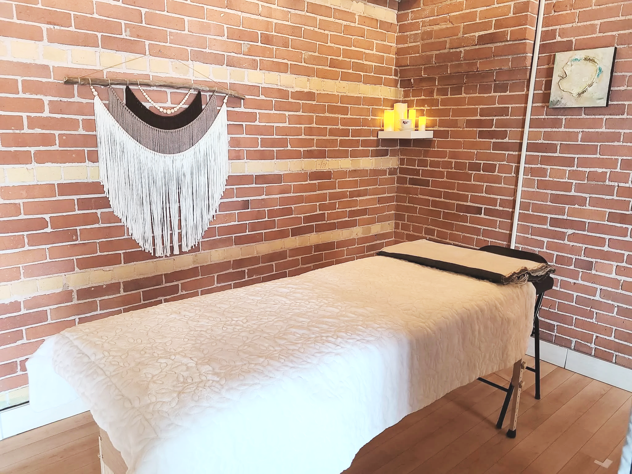 Interior of a room with exposed red brick walls. There is a bed with a light-colored quilt, a small folding chair next to it, a wall shelf with candles, a wall hanging with black, white, and bead details, and a piece of abstract art on the wall.