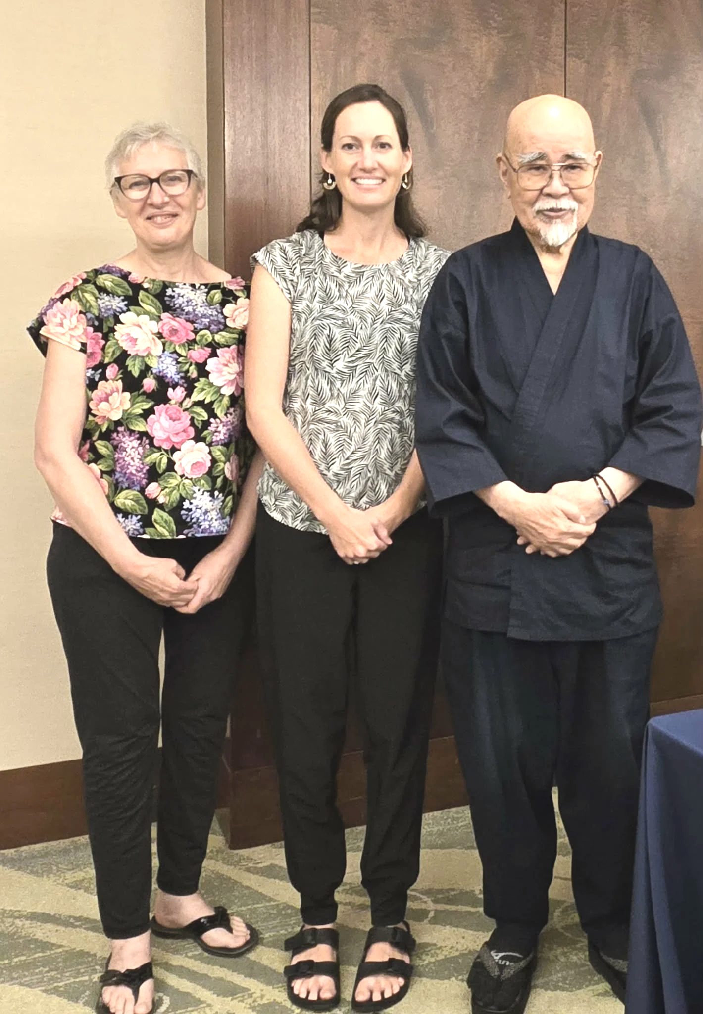 Three people standing indoors in front of a wooden wall. The person on the left is a woman with short blonde hair, glasses, a floral top, black pants, and sandals. The person in the middle is a woman with shoulder-length dark hair, wearing earrings, a patterned top, black pants, and sandals. The person on the right is a man with a bald head and a white beard, dressed in traditional black martial arts attire, and black shoes.