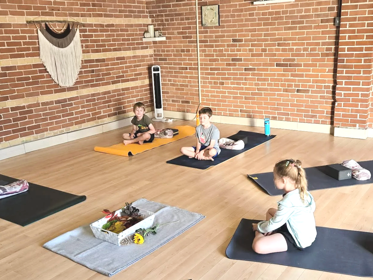 Three children sitting on yoga mats in a yoga studio with brick walls, practicing meditation or mindfulness, with a decorative wall hanging and a basket with flowers and leaves in the foreground.