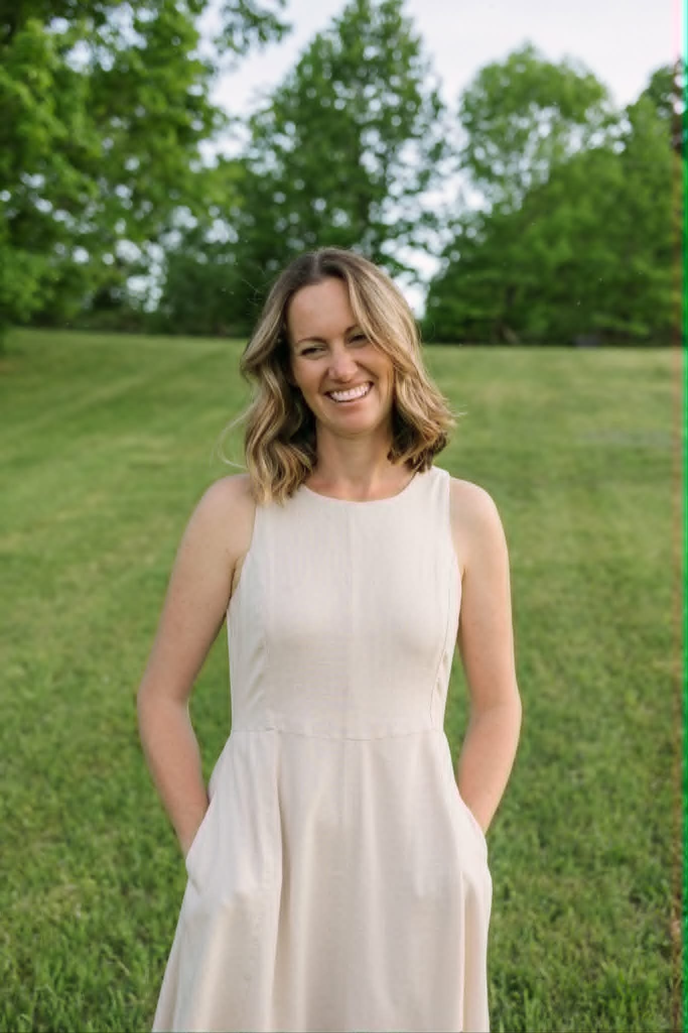 A woman with shoulder-length light brown hair wearing a sleeveless cream-colored dress standing on a grassy field with green trees in the background, smiling.