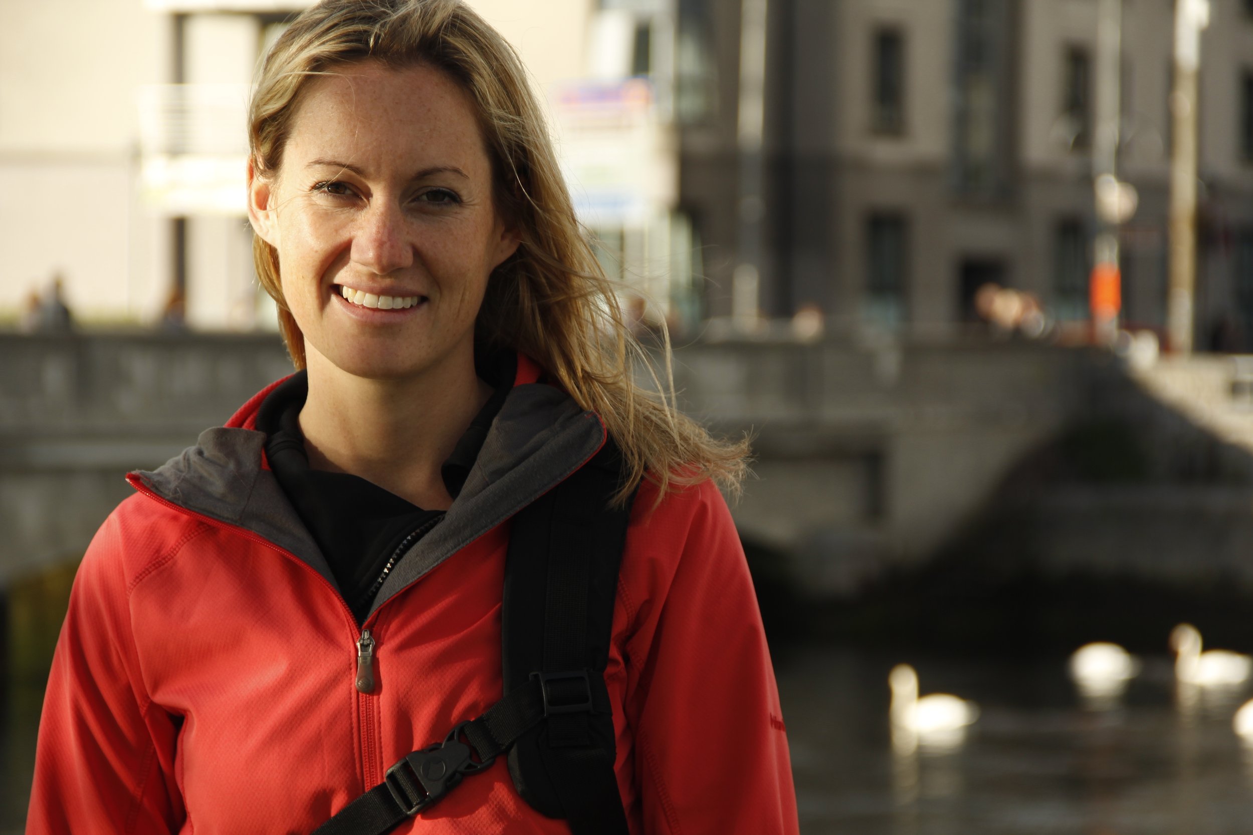 A woman with red hair and freckles smiling outdoors in urban area, wearing a red jacket and black backpack strap.