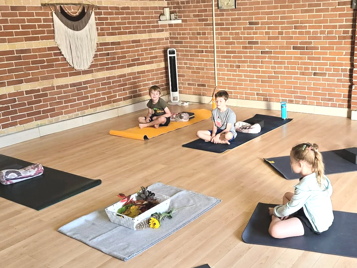 Children doing yoga in a studio with brick walls, mats, and a decorative wall hanging.