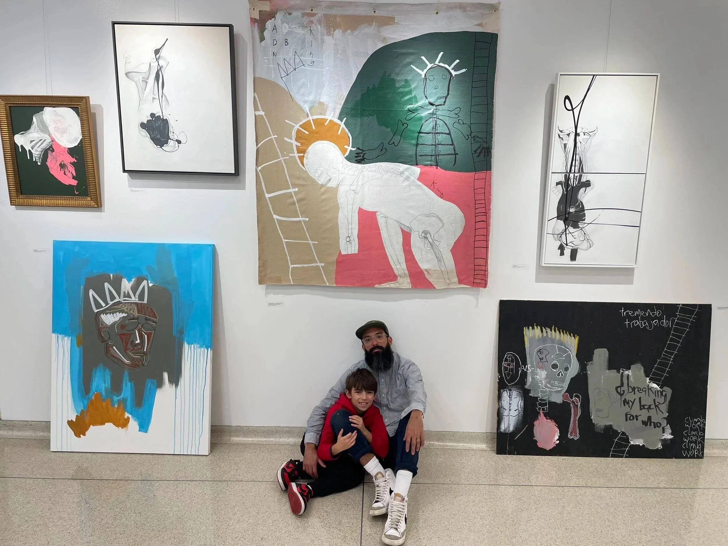 A man and a young boy sitting and smiling in front of colorful abstract artwork at an art gallery.