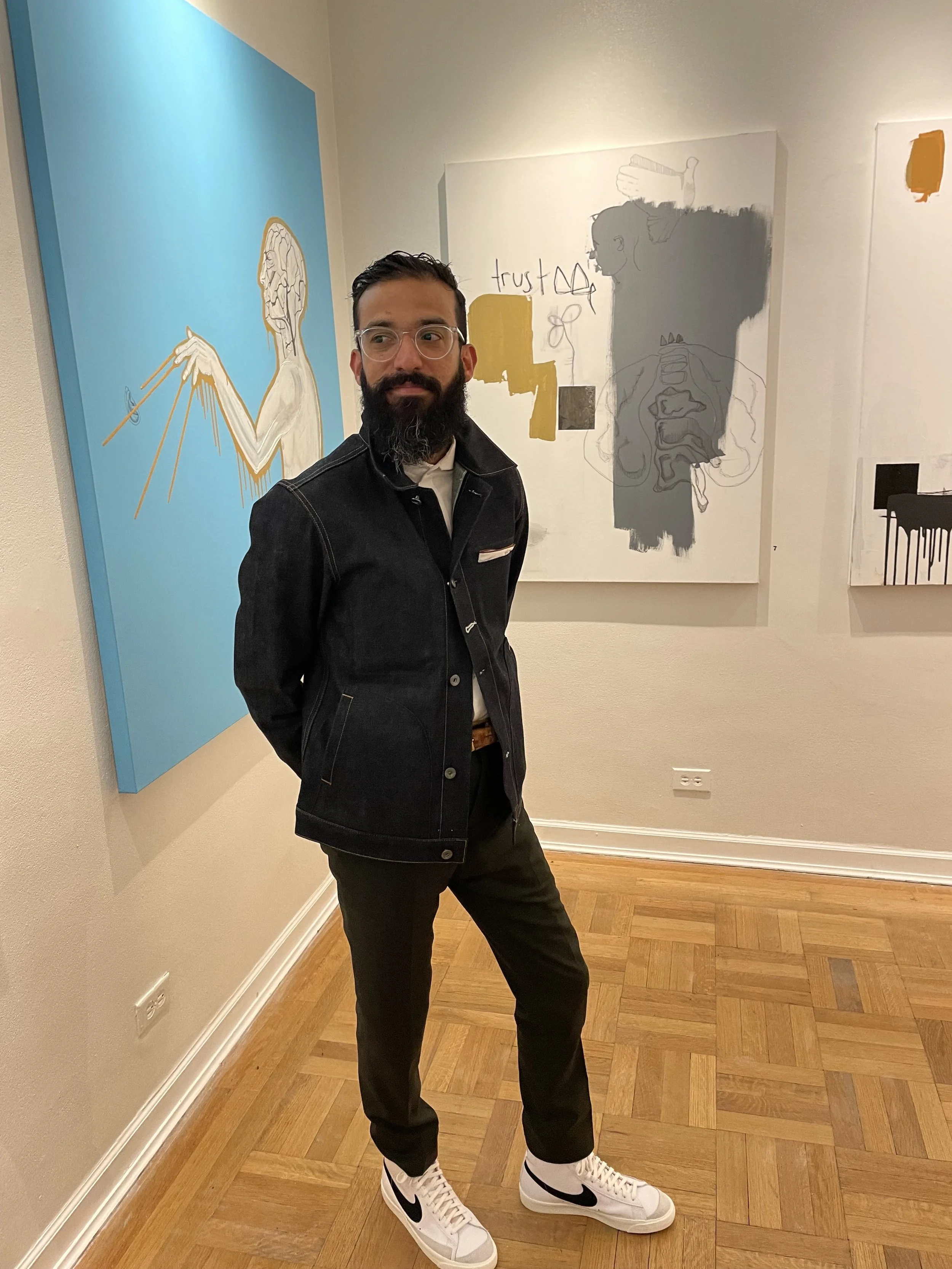 A man with glasses and a beard standing in an art gallery, wearing black pants, a jacket, and white Nike sneakers, with colorful artworks on the wall behind him.