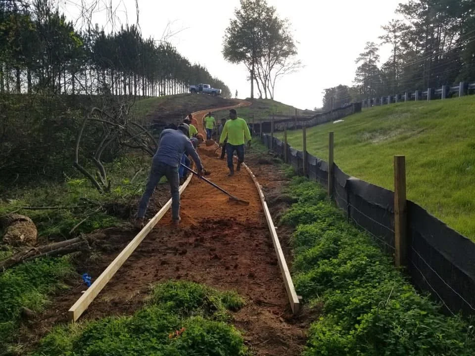 Construction workers installing a gravel or dirt pathway along a hill, with several people working and a vehicle at the top of the hill.