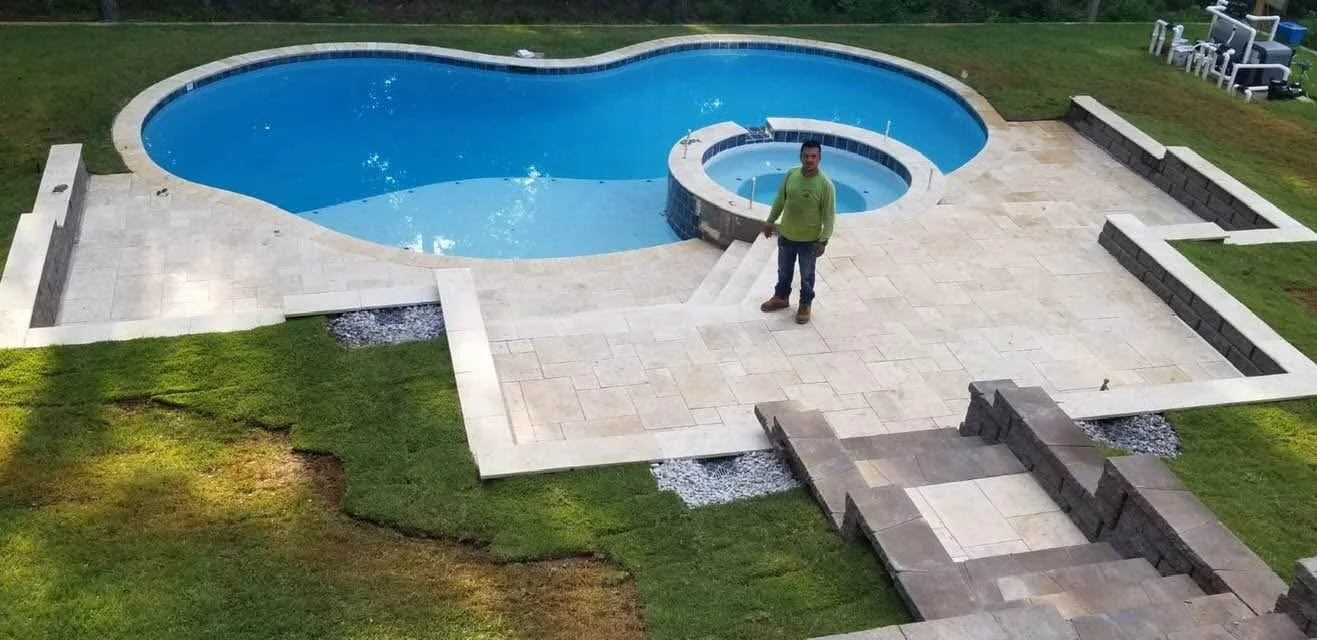 Man standing by a backyard swimming pool with a large tiled patio, surrounded by grass and garden beds.