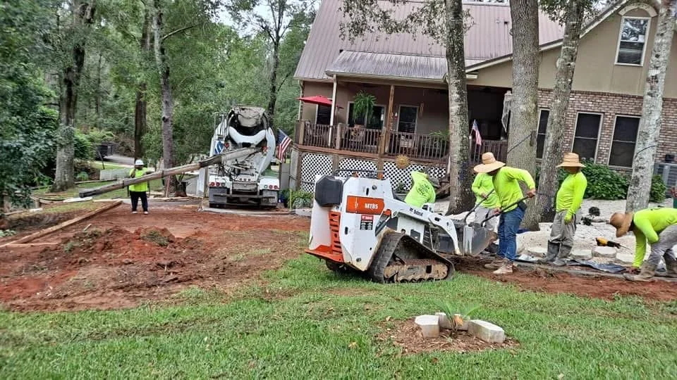Workers installing or repairing a driveway with heavy machinery near a house, surrounded by trees.