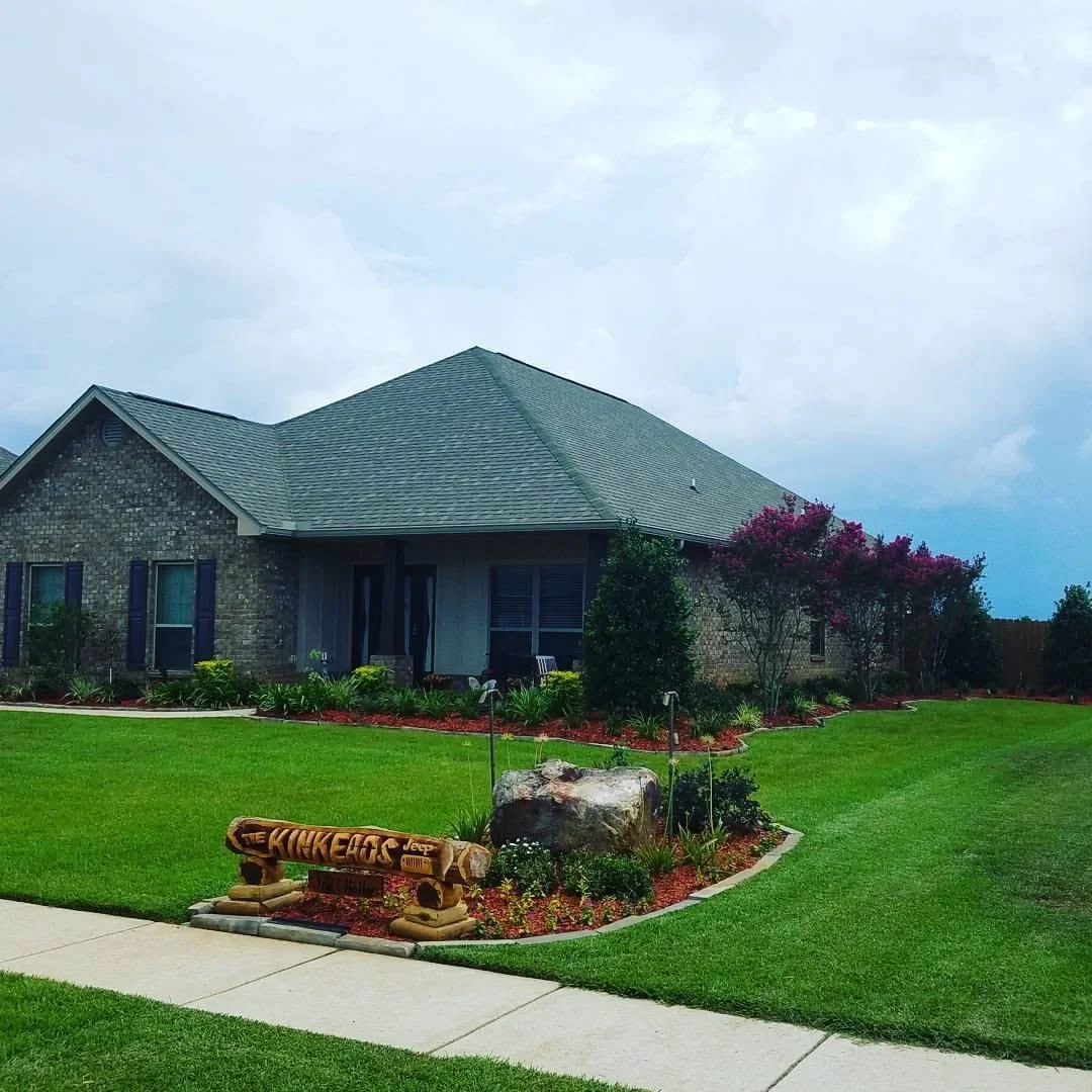 A house with brick and siding exterior, surrounded by a well-maintained lawn and flower beds, with a rock and a wooden bench that has the Nike logo on it in the front yard.