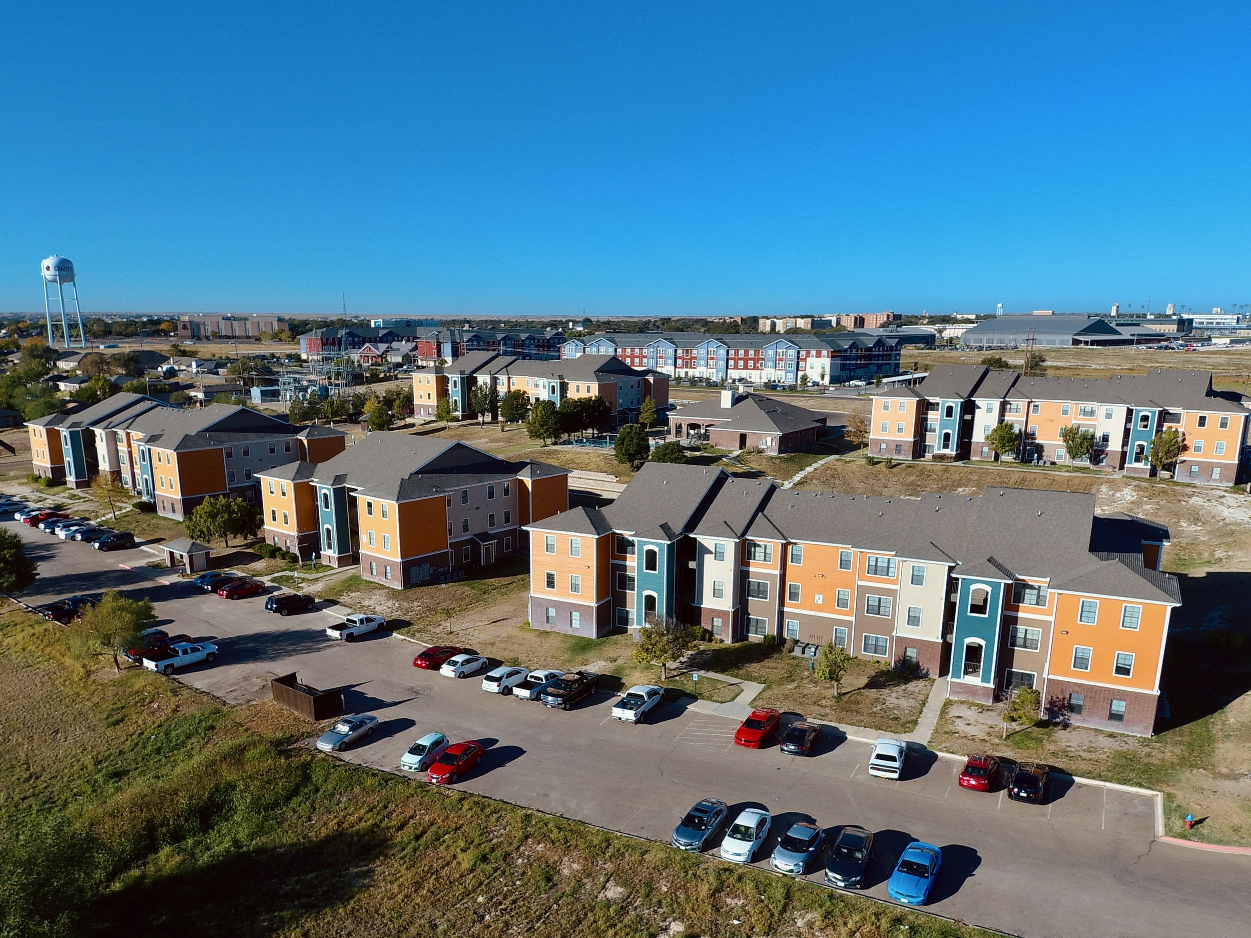 Aerial view of a residential area with colorful apartment buildings, parking lots with cars, a water tower, and industrial buildings in the background under a clear blue sky.