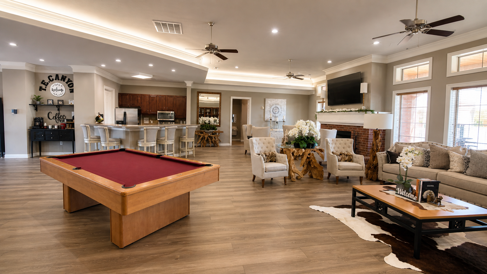 Upscale living room with pool table, beige armchairs, floral arrangements, large flat-screen TV, fireplace, and kitchen area with bar stools.