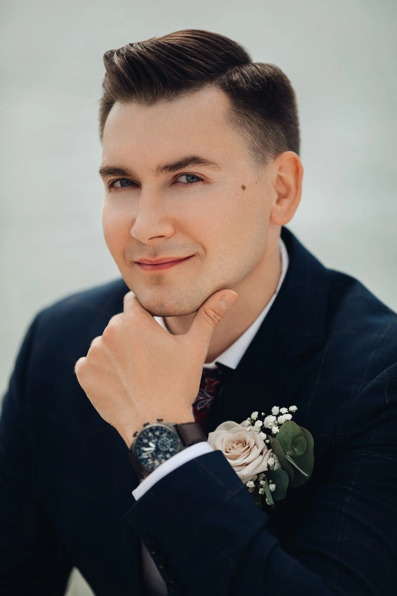 Close-up of a groom in a navy blue suit, white shirt, and floral tie, with a boutonniere of white roses and greenery, posing with his hand on his chin.