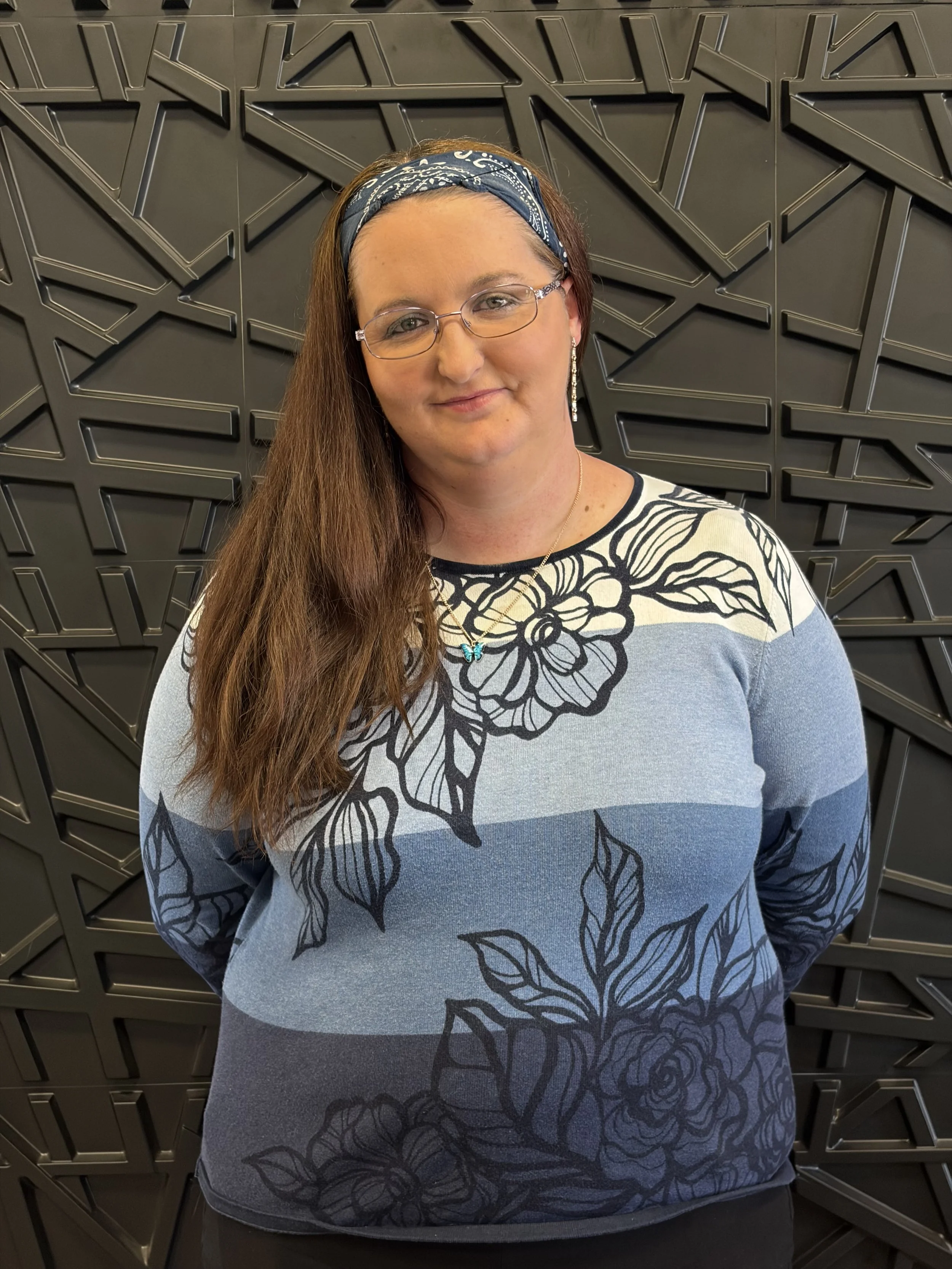 A woman with glasses and a blue bandana headband smiling, standing in front of a dark textured wall with geometric patterns, wearing a floral patterned sweater.