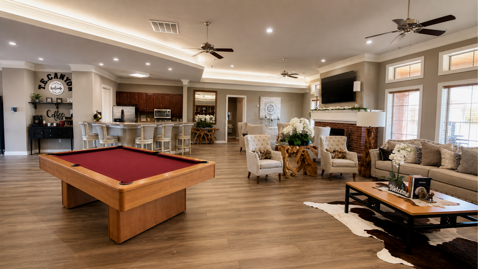 Spacious living room with a pool table, beige armchairs, a sofa, a fireplace, a large flat-screen TV, and a bar area with barstools. Large windows letting in natural light, wood flooring, and decorative plants and flowers.