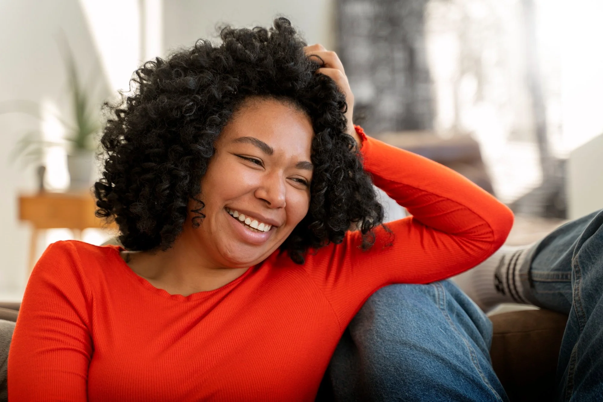 A woman with curly black hair smiling and laughing, wearing an orange top, sitting on a couch with a person in jeans and socks nearby.