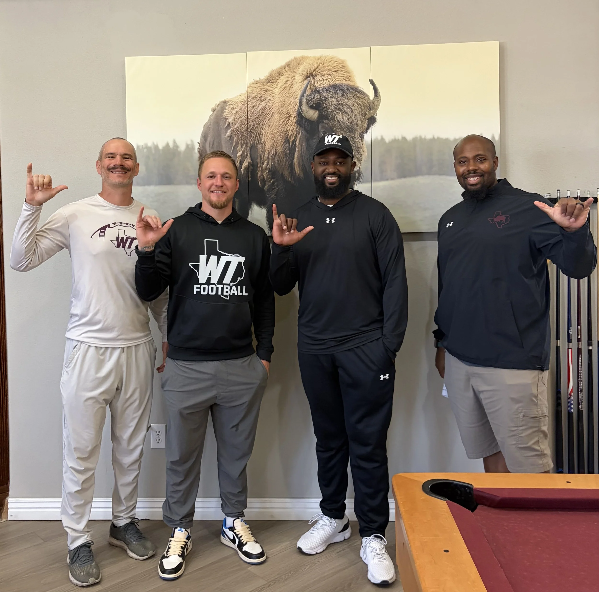 Four men standing in front of a wall with a large photo of a bison. They are all smiling and making hand gestures. The man on the far left is wearing a white long-sleeve shirt and tan pants, the second man is in a black hoodie and gray pants, the third man is in a black hoodie and dark pants, and the man on the far right is wearing a black jacket and tan pants.