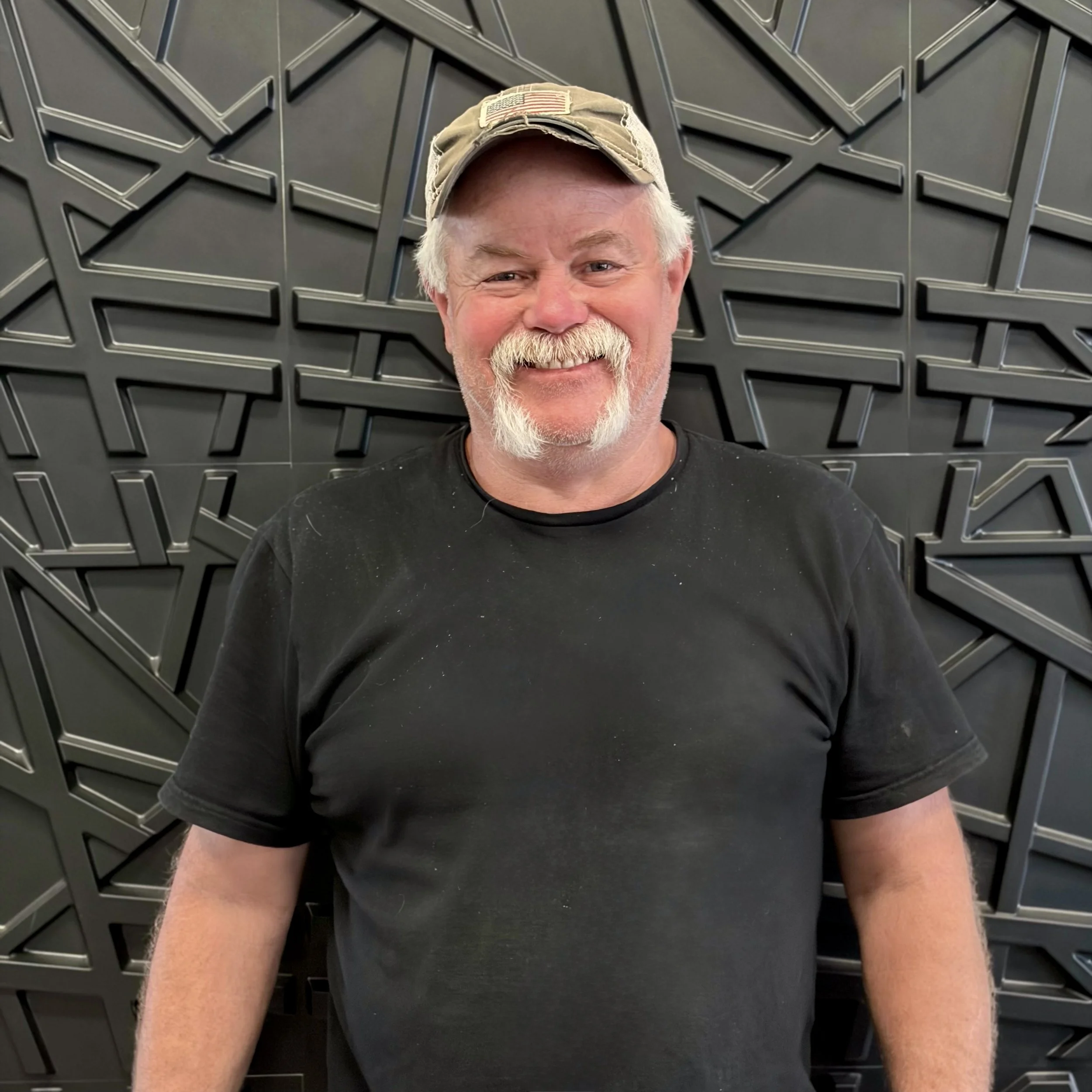 A man with a white beard and mustache wearing a camouflage baseball cap with an American flag patch, smiling at the camera, standing in front of a textured black background.