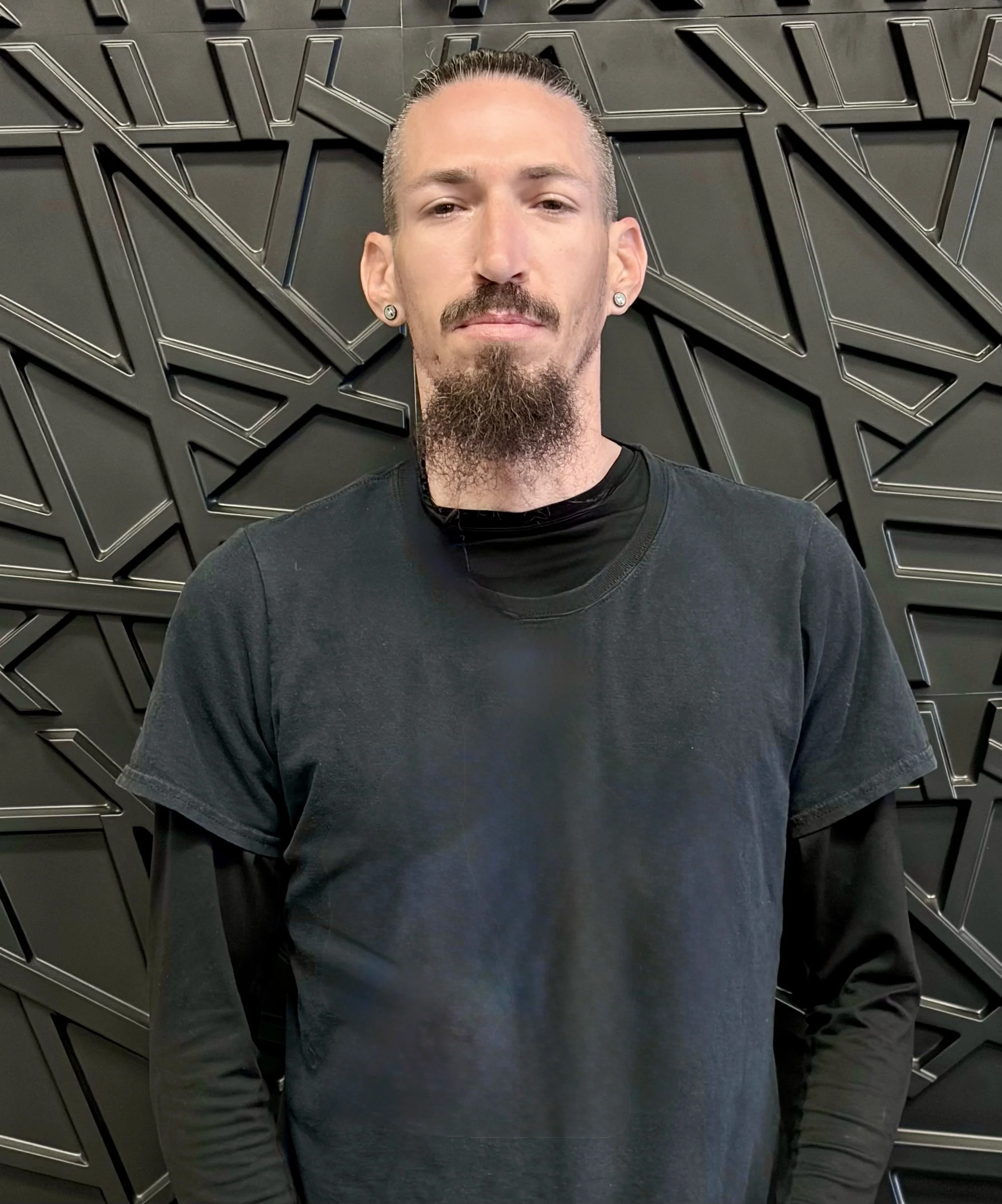 A man with a goatee and earrings standing in front of a geometric, black, metal wall design.