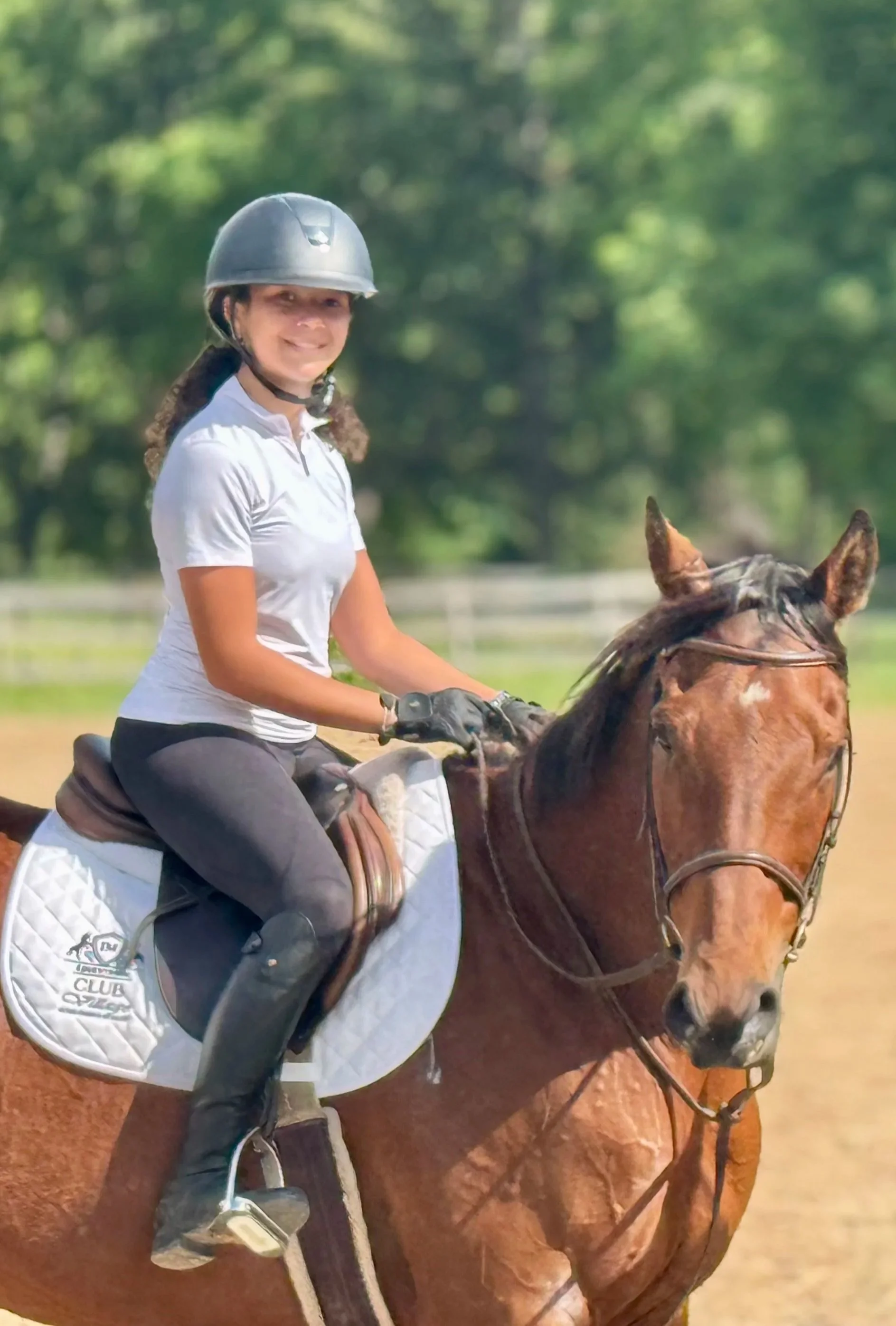 A woman wearing a riding helmet and gloves, smiling while riding a horse outdoors on a sunny day with trees in the background.