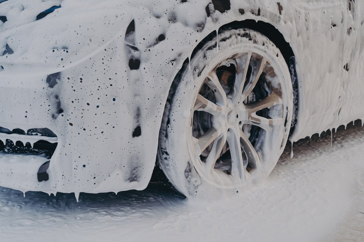 A car being washed with white soap suds covering the front left wheel and part of the car body, with foam dripping down.