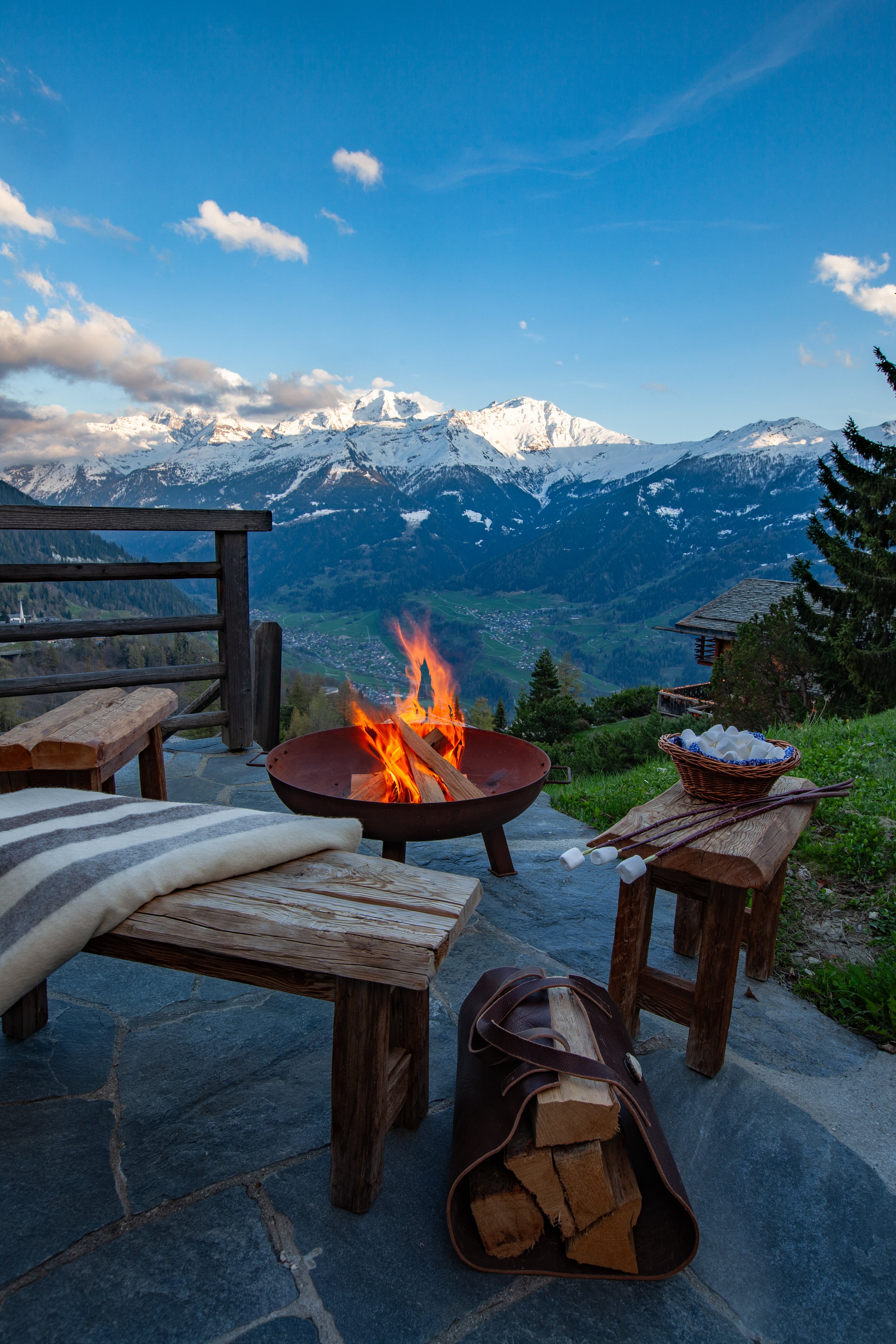 Outdoor fire pit at dusk with handmade wooden benches and panoramic Alpine views, Chalet la Vigne Verbier Switzerland