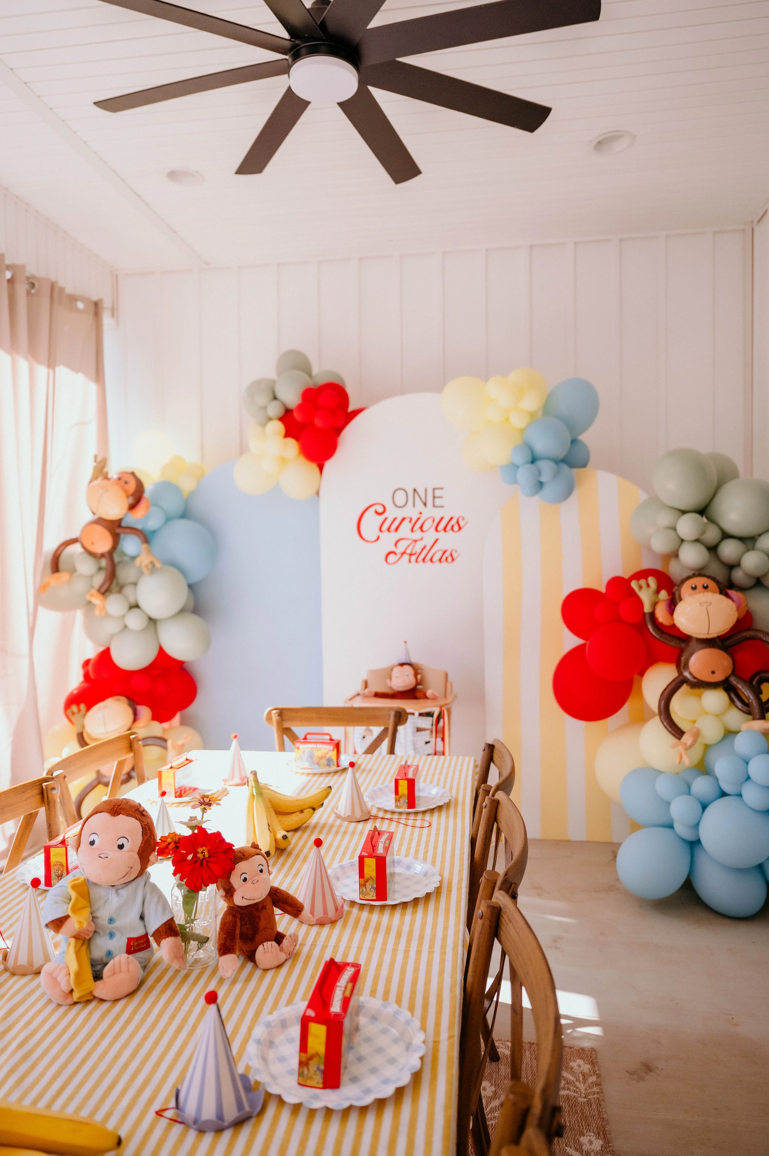 A decorated party table with stuffed monkey toys, party hats, red boxes, and a birthday banner that reads "One Curious Atlas." The room features balloon decorations in pastel colors and a ceiling fan.