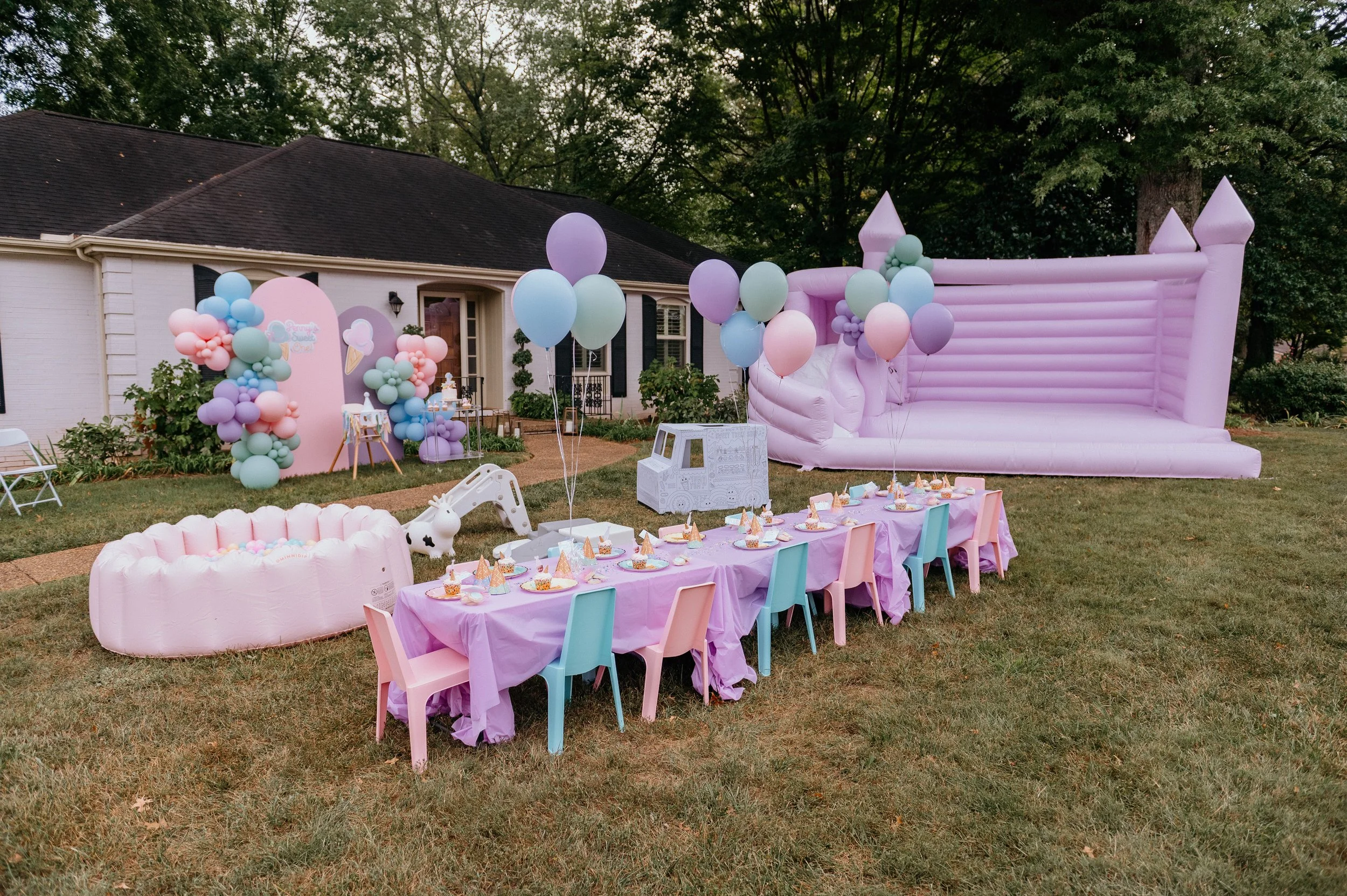 Decorated backyard with pink and pastel-colored balloons, a pink inflatable castle, a table set for a celebration with party hats and cupcakes, and a teepee with a unicorn figure, preparing for a children's birthday party.