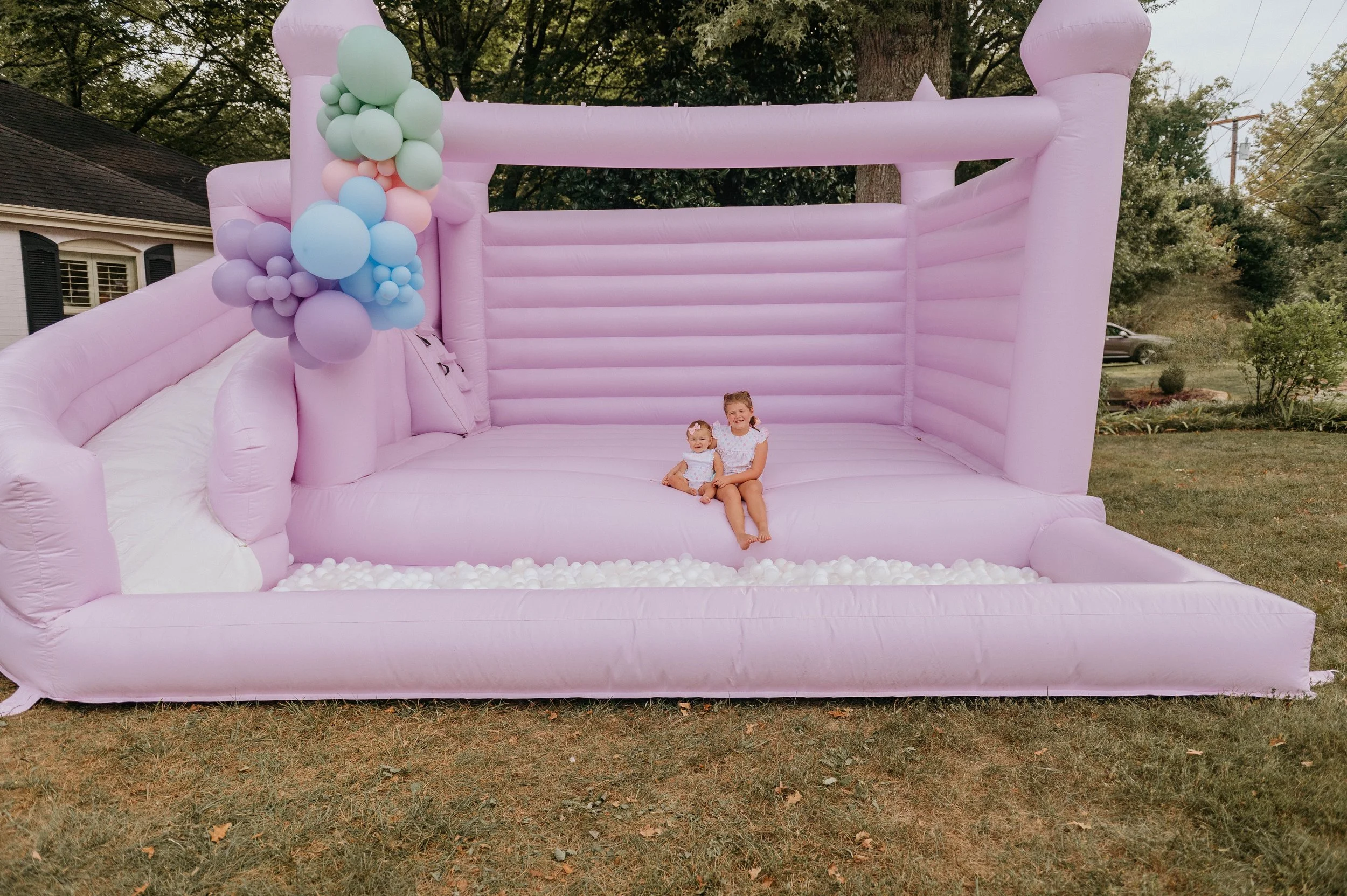 A pink inflatable bounce house with a ball pit containing white balls, decorated with pastel-colored balloons on the left side. Two young girls are sitting inside, smiling.