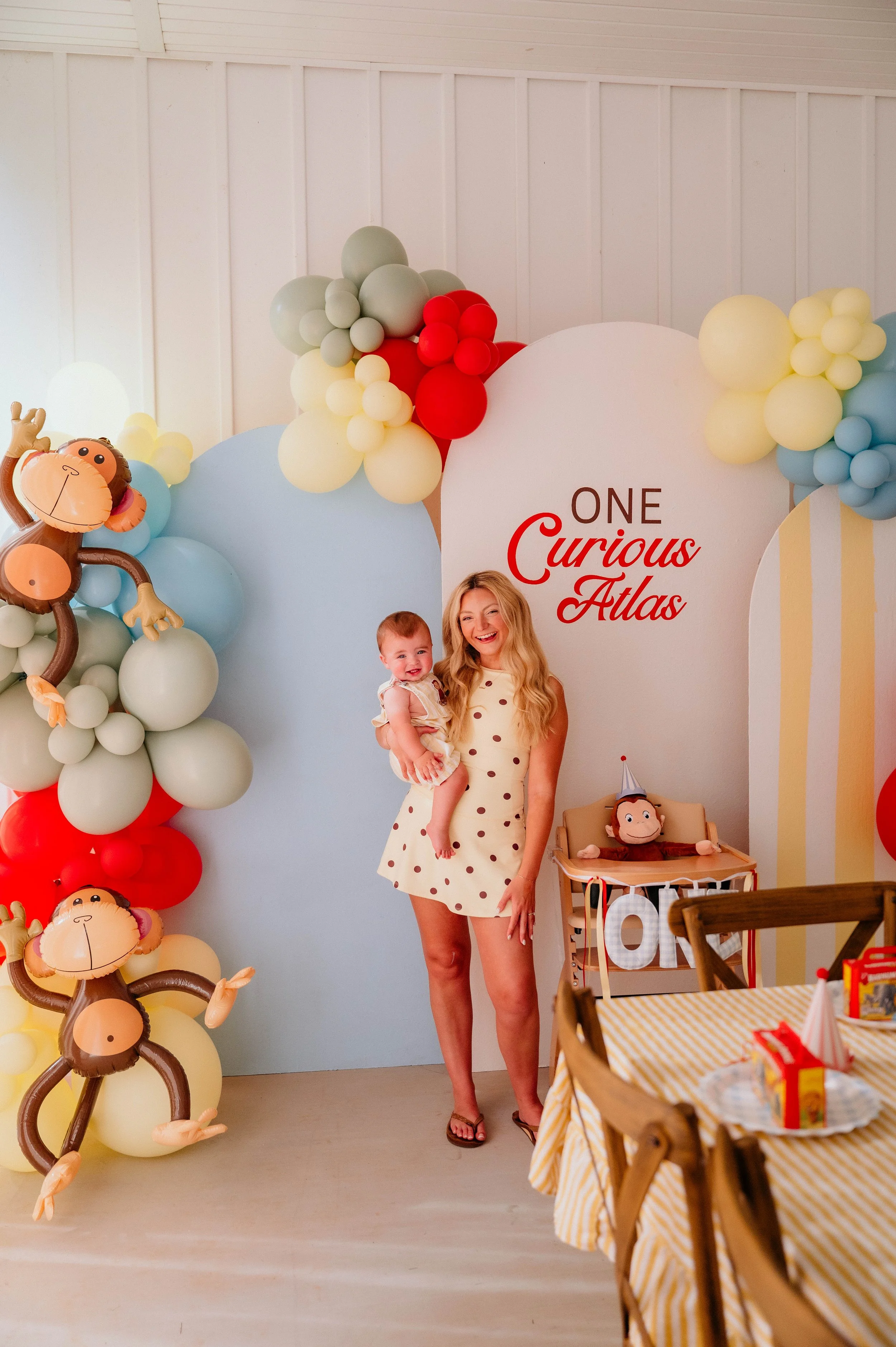 A woman holding a child at a birthday party with monkey-themed balloons and a backdrop that reads 'One Curious Atlas'.