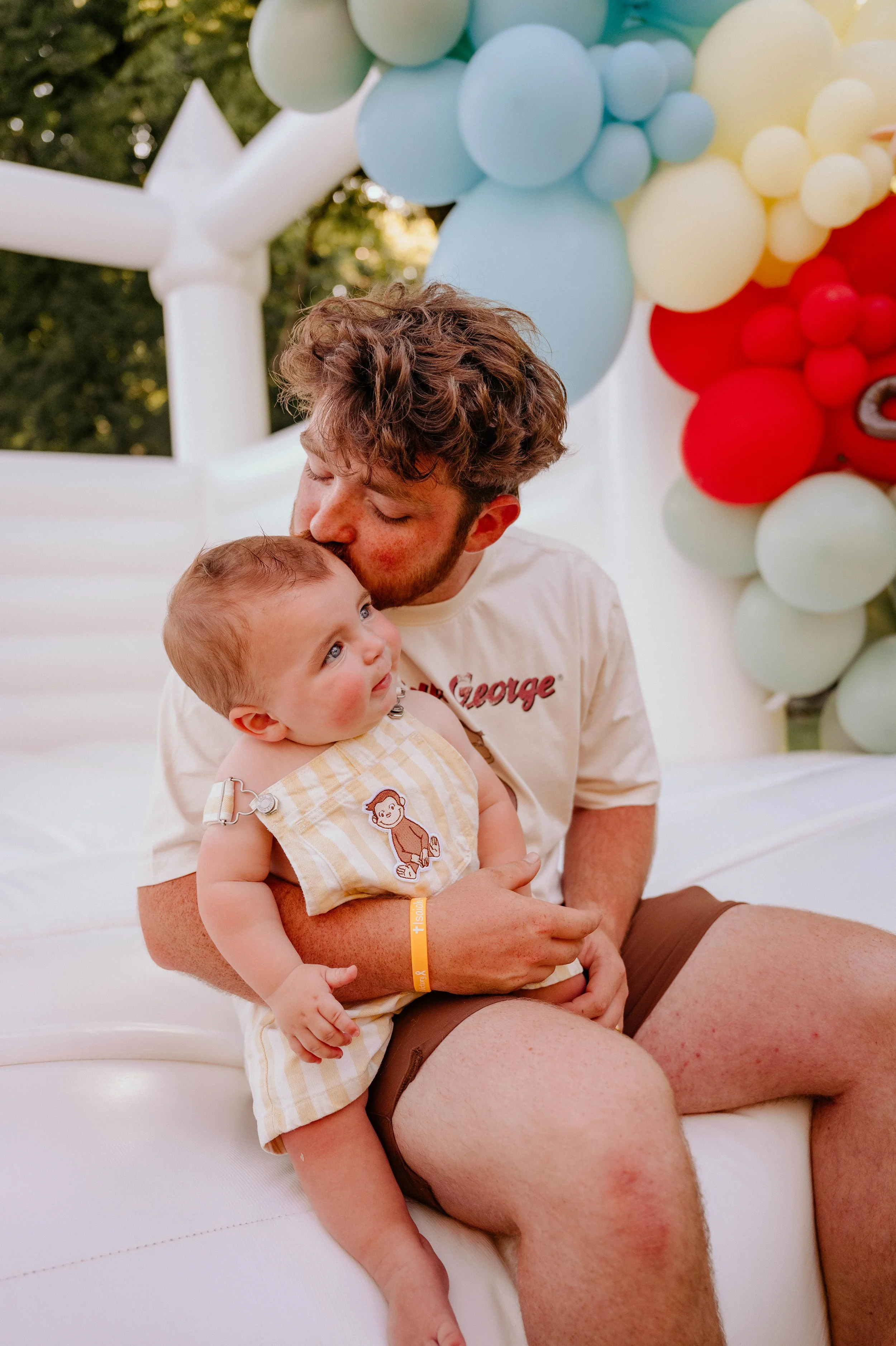 A man with light skin and brown, curly hair is holding a young child with light skin and blonde hair, both sitting on a white surface outdoors. The background features a colorful balloon decoration with red, blue, yellow, and cream balloons.