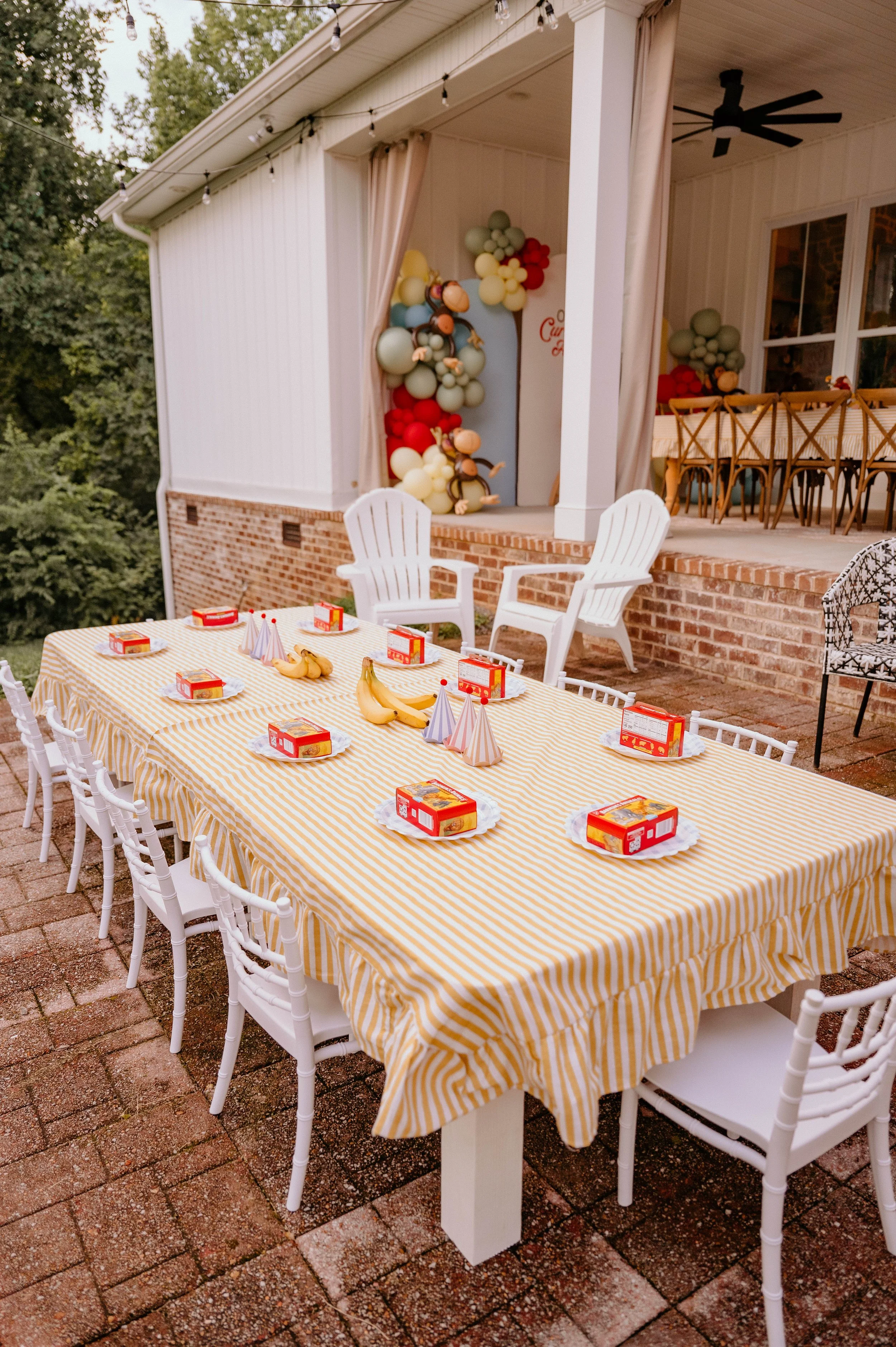 Outdoor birthday party setup with a long table covered in a yellow and white striped tablecloth, decorated with bananas, party hats, and juice boxes, surrounded by white chairs. In the background, there is a porch with a Christmas scene made of ballo