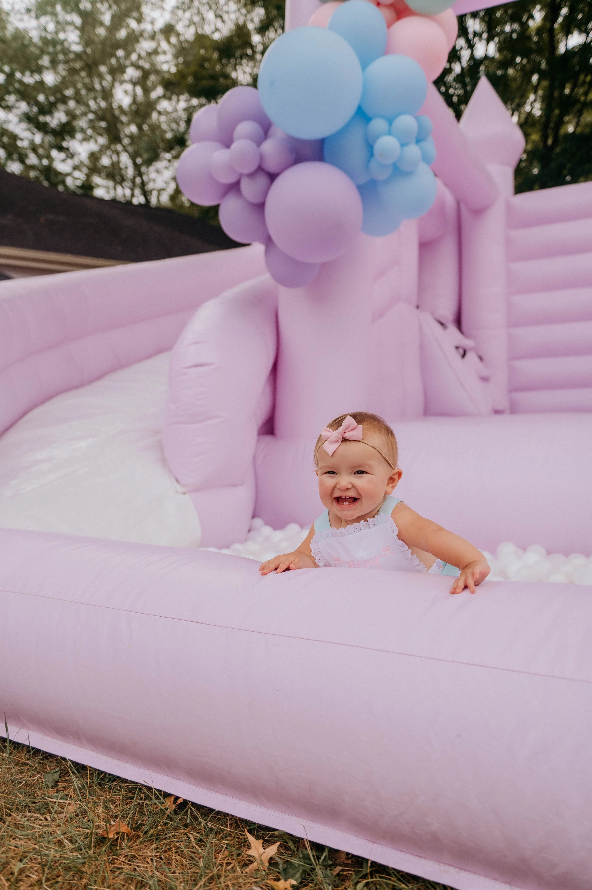 A baby girl in a white dress with pink bow headband playing on a pink inflatable bounce house decorated with pastel-colored balloons, outdoors on grass.