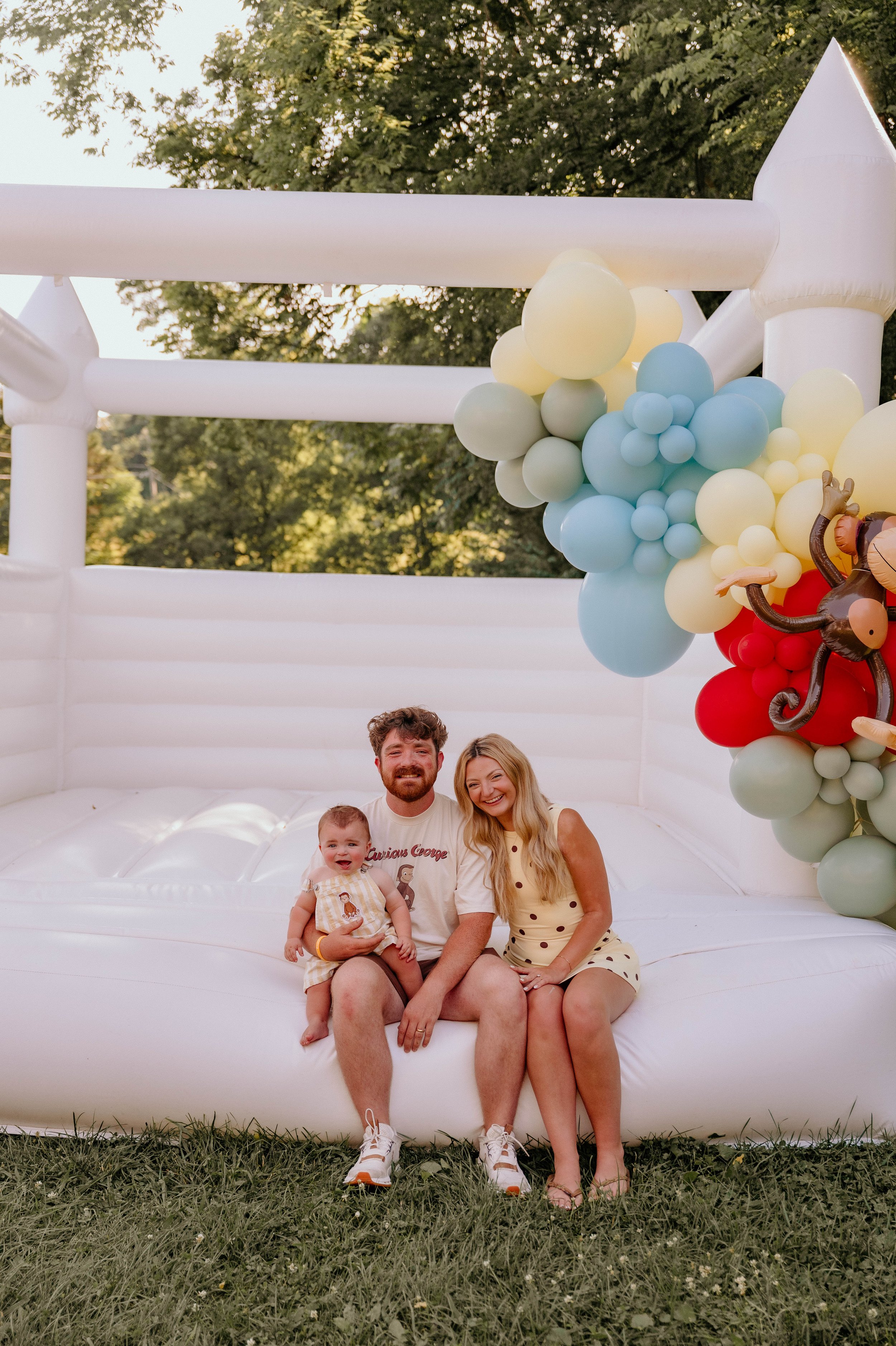 A family of four sitting on an inflatable white structure with colorful balloon decorations in an outdoor setting, surrounded by trees and grass.
