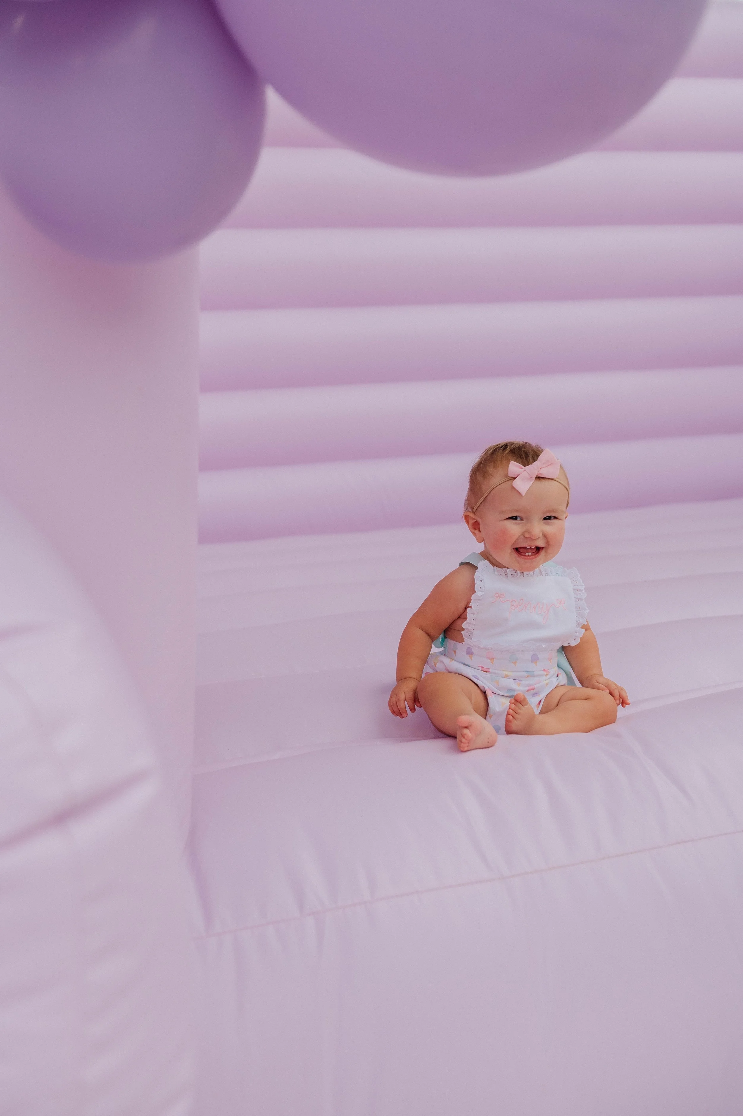 A smiling baby girl with a pink bow headband sitting on a pink inflatable bounce house, wearing a white sleeveless outfit with frills.
