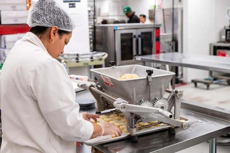A worker in a commercial kitchen using a machine to shape dough into small cookies or pastries, with other kitchen staff and equipment visible in the background.