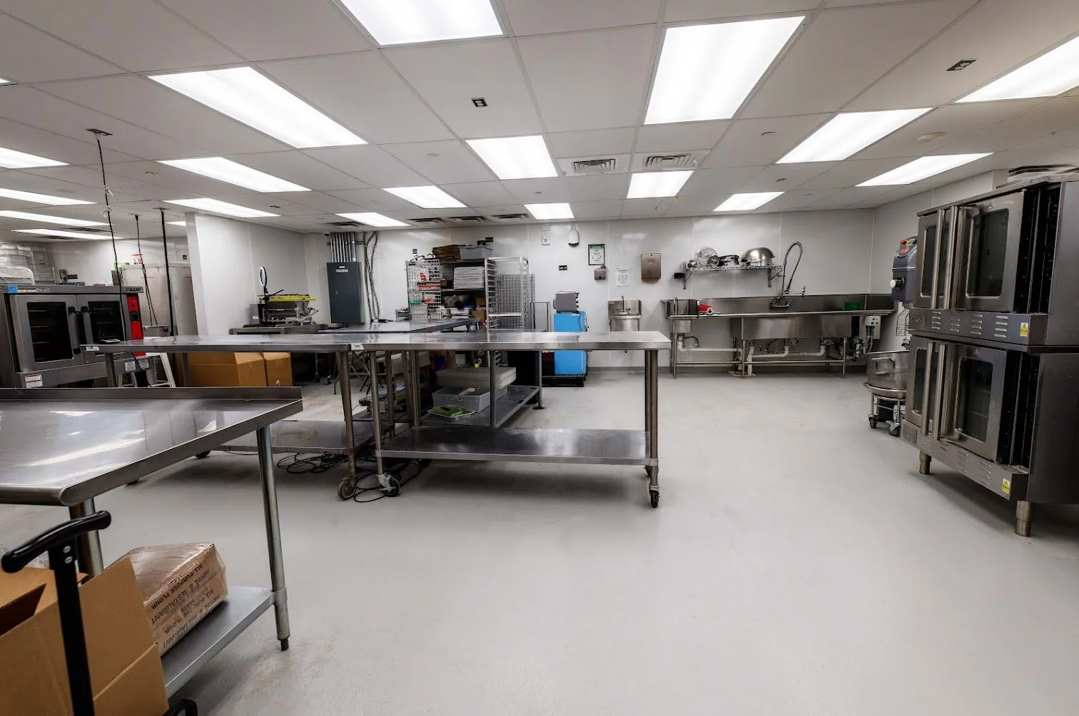 An empty commercial kitchen with stainless steel tables, industrial ovens, racks, and cleaning stations, illuminated by bright ceiling lights.