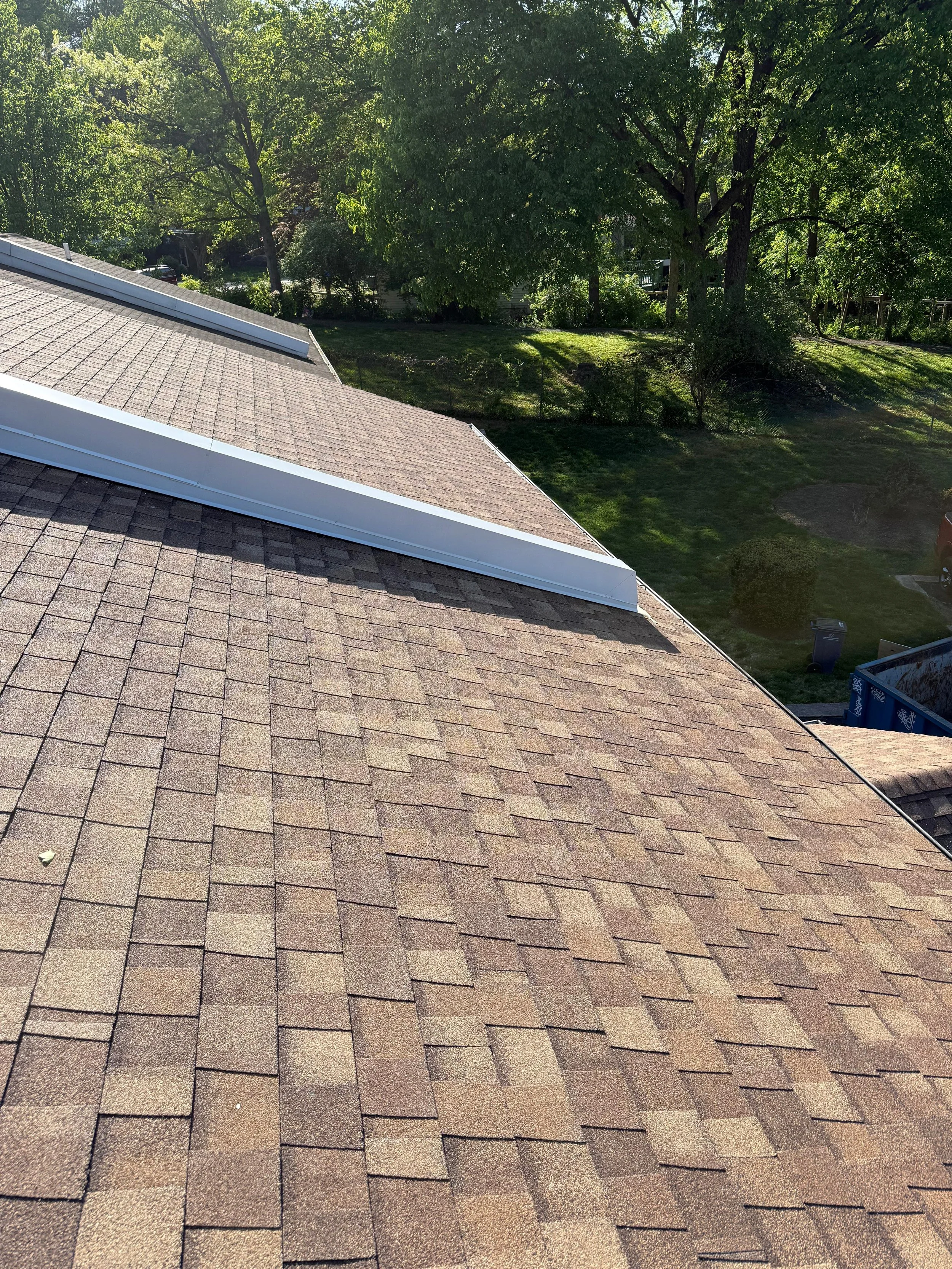 View of a residential roof with asphalt shingles, surrounded by green trees and a yard, captured in bright sunlight.